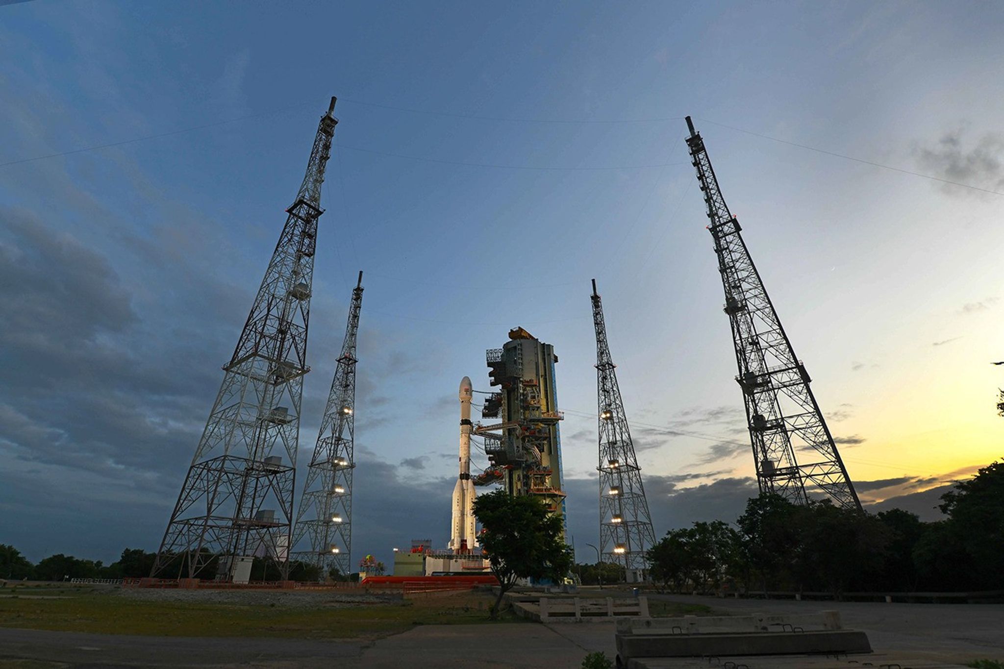 A wide, low-angle shot of a rocket on a launch pad at dusk. The white rocket is positioned next to its mobile service tower and is surrounded by four tall, illuminated metal lattice towers. The sky behind is a deep blue with clouds, transitioning to a warm yellow and orange glow from the setting sun on the right horizon.