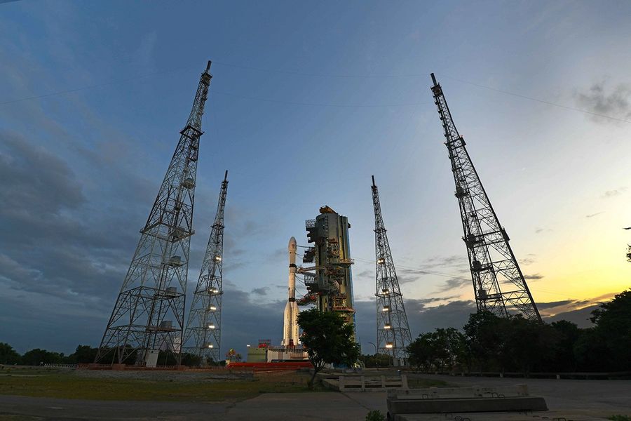A wide, low-angle shot of a rocket on a launch pad at dusk. The white rocket is positioned next to its mobile service tower and is surrounded by four tall, illuminated metal lattice towers. The sky behind is a deep blue with clouds, transitioning to a warm yellow and orange glow from the setting sun on the right horizon.