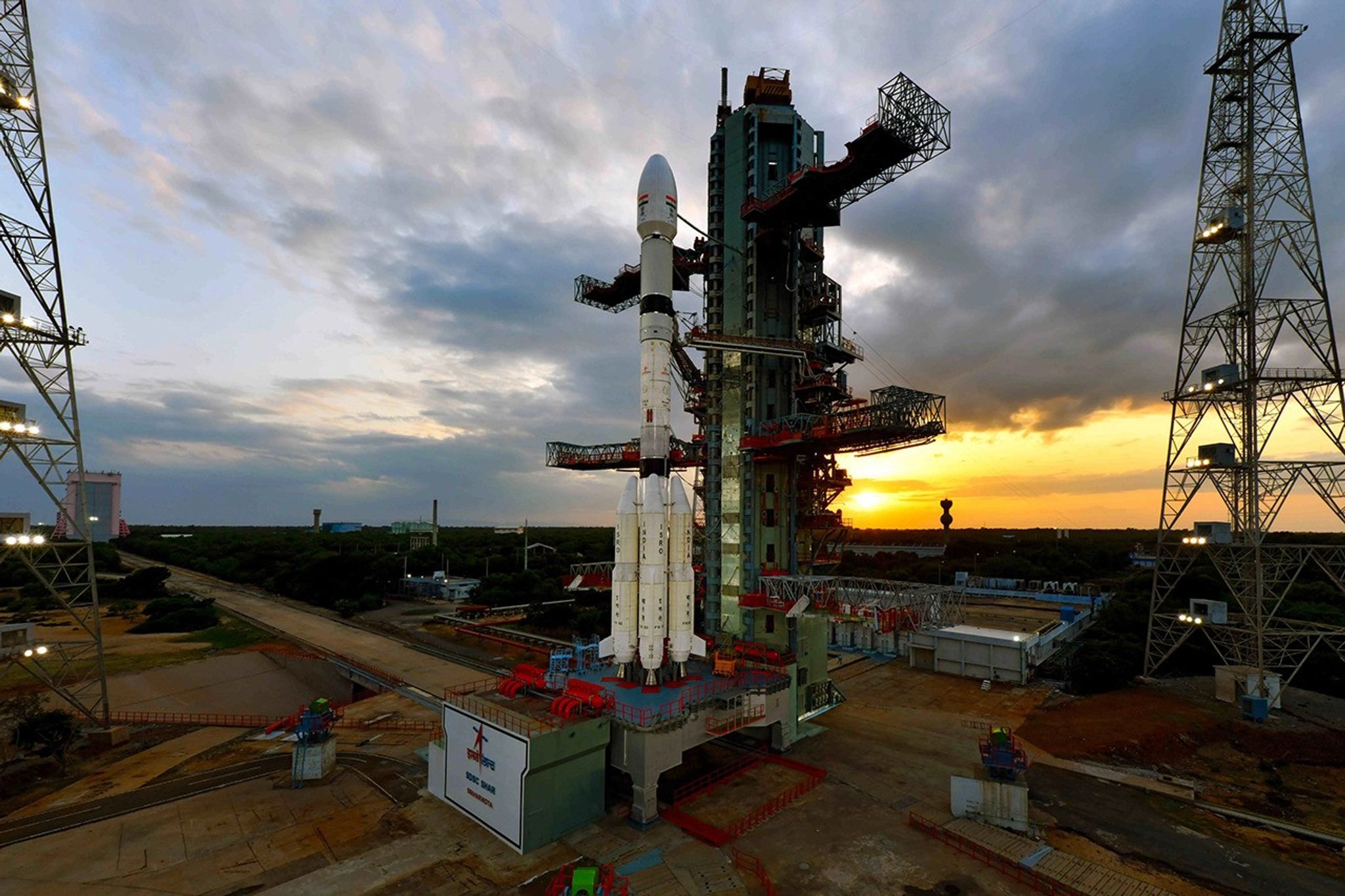 An elevated, wide photograph of a rocket on its launchpad. The white rocket, with booster rockets attached, stands vertically next to its large, multi-level service tower. The launch complex is surrounded by green vegetation, and in the background, a dramatic, cloudy sky is illuminated by the vibrant orange and yellow glow of a sunrise or sunset on the horizon.