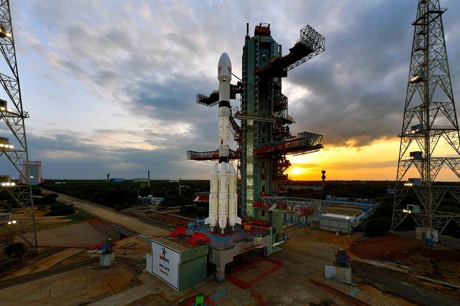An elevated, wide photograph of a rocket on its launchpad. The white rocket, with booster rockets attached, stands vertically next to its large, multi-level service tower. The launch complex is surrounded by green vegetation, and in the background, a dramatic, cloudy sky is illuminated by the vibrant orange and yellow glow of a sunrise or sunset on the horizon.