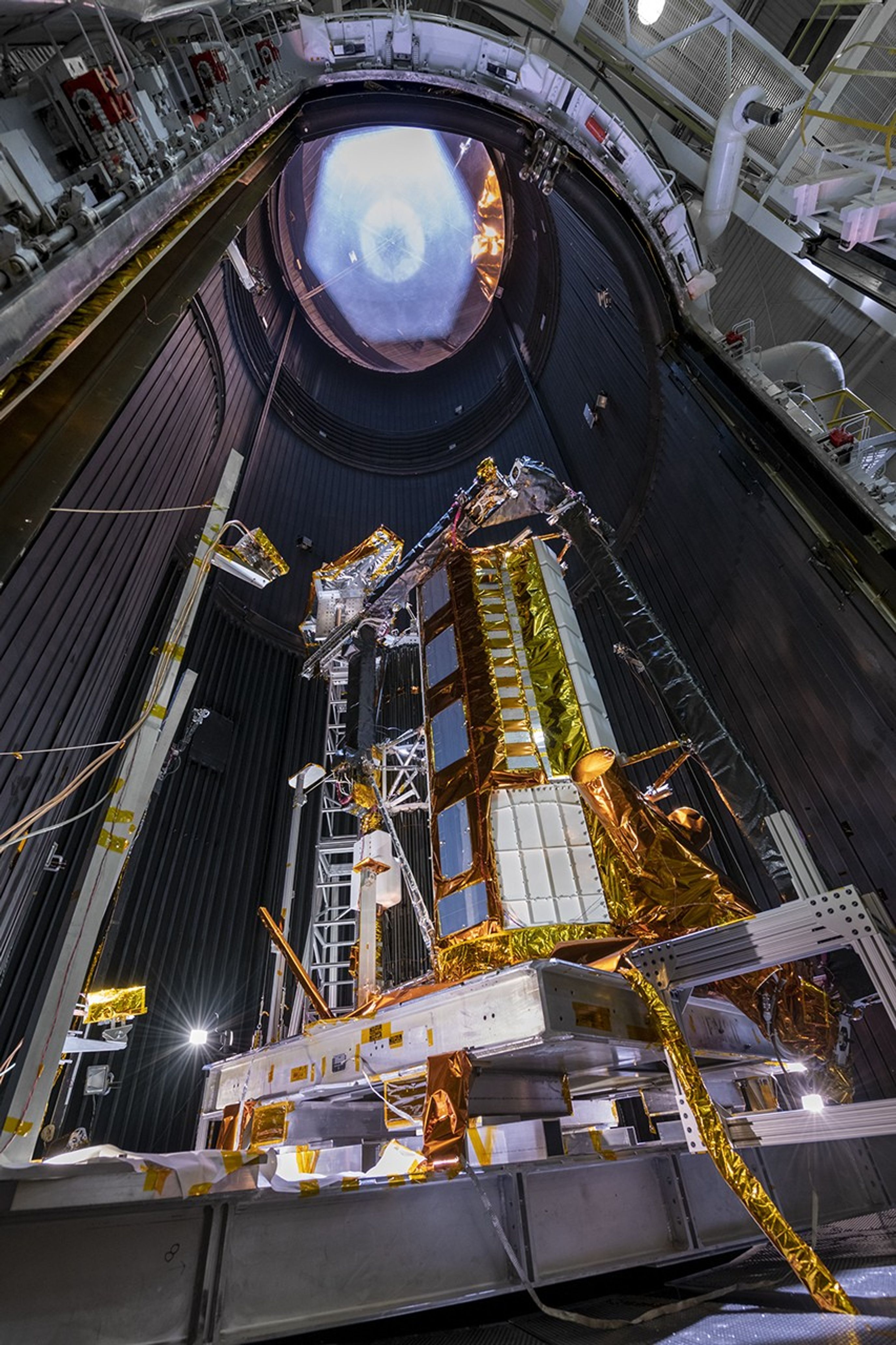 NISAR's flight antenna system stands in the center of a thermal vacuum chamber at NASA's Jet Propulsion Laboratory. The image is taken at the base of the structure, and the antenna looms over the frame from this point of view, revealing its impressive size.