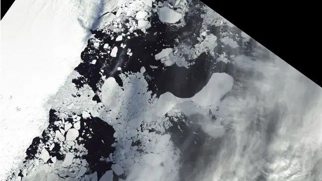 Along the left side of the image is completely ice covered solid mass. the remainder of the image is a sporadic mix of white ice and near black ocean. Clouds obscuring the rightmost edge of the image.