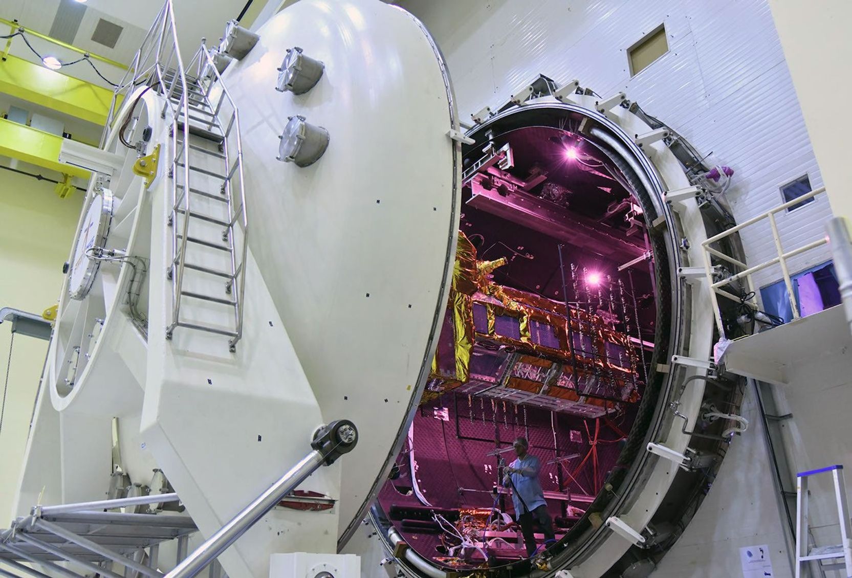 a large white structure inside of a white building appears open with a purple interior and what appears to be the gold covered NISAR satellite.
