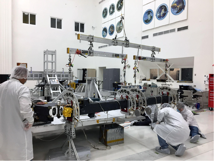 Engineers crouch around NISAR's upper antenna boom, inspecting it before assembly. The hardware hangs from three metal bars to keep it elevated for easy inspection.