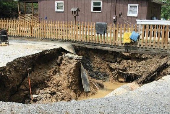 A sinkhole jeopardizes a nearby home's structural integrity. The brick of the basement is visible and damaged. The yard and fence above hold together despite the gravel road's collapse.