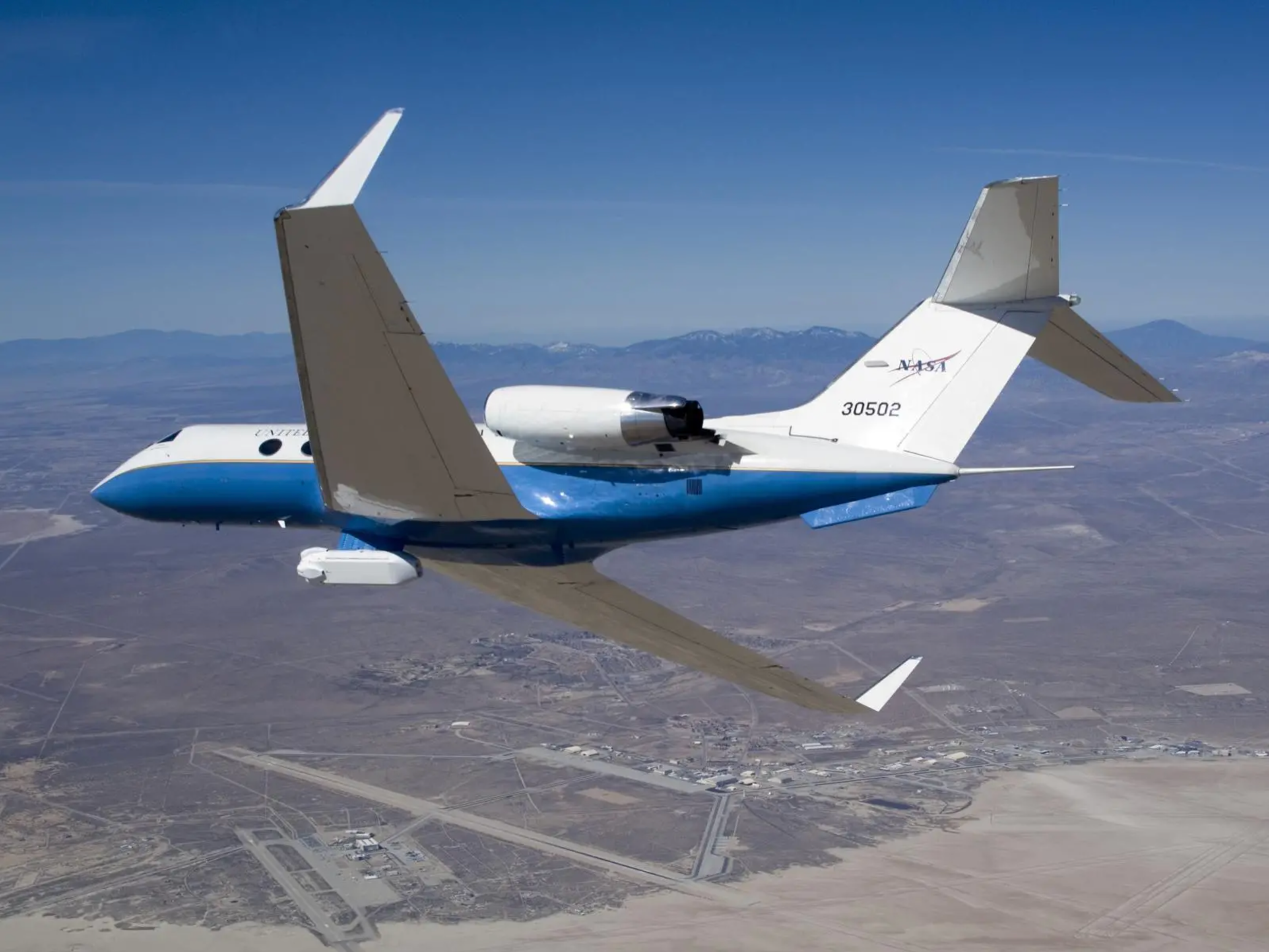 Gulfstream plane with NASA logo flies over arid landscape. A mountain range is in the distance near the horizon.