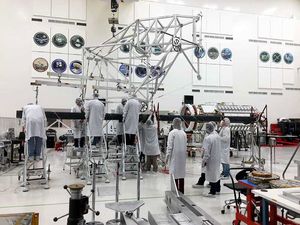 After mating the upper and lower boom assemblies, engineers at NASA's Jet Propulsion Laboratory inspect the fully deployed structure for alignment. A team of 11 individuals in white coats and hairnets huddle around the structure.