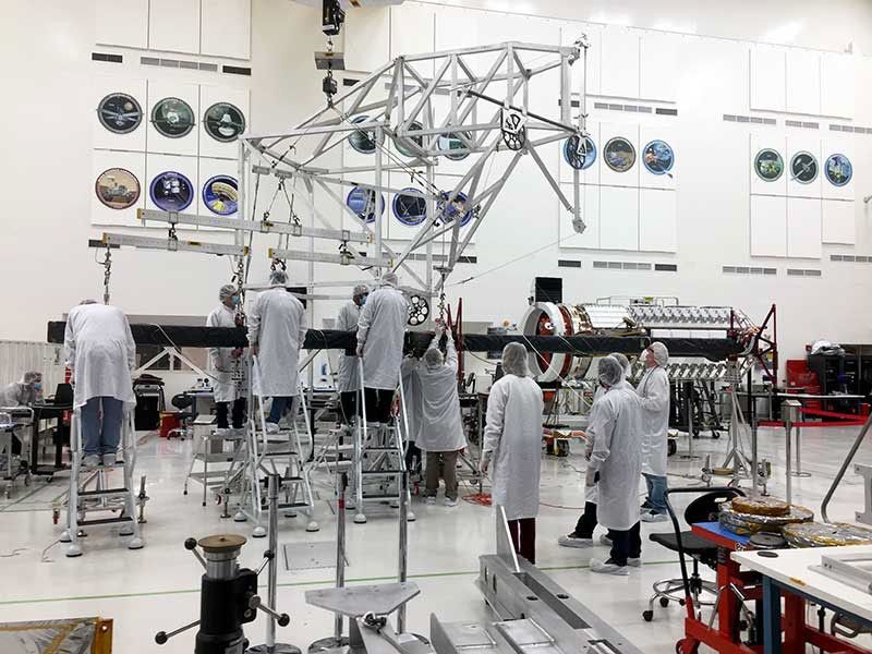 After mating the upper and lower boom assemblies, engineers at NASA's Jet Propulsion Laboratory inspect the fully deployed structure for alignment. A team of 11 individuals in white coats and hairnets huddle around the structure.