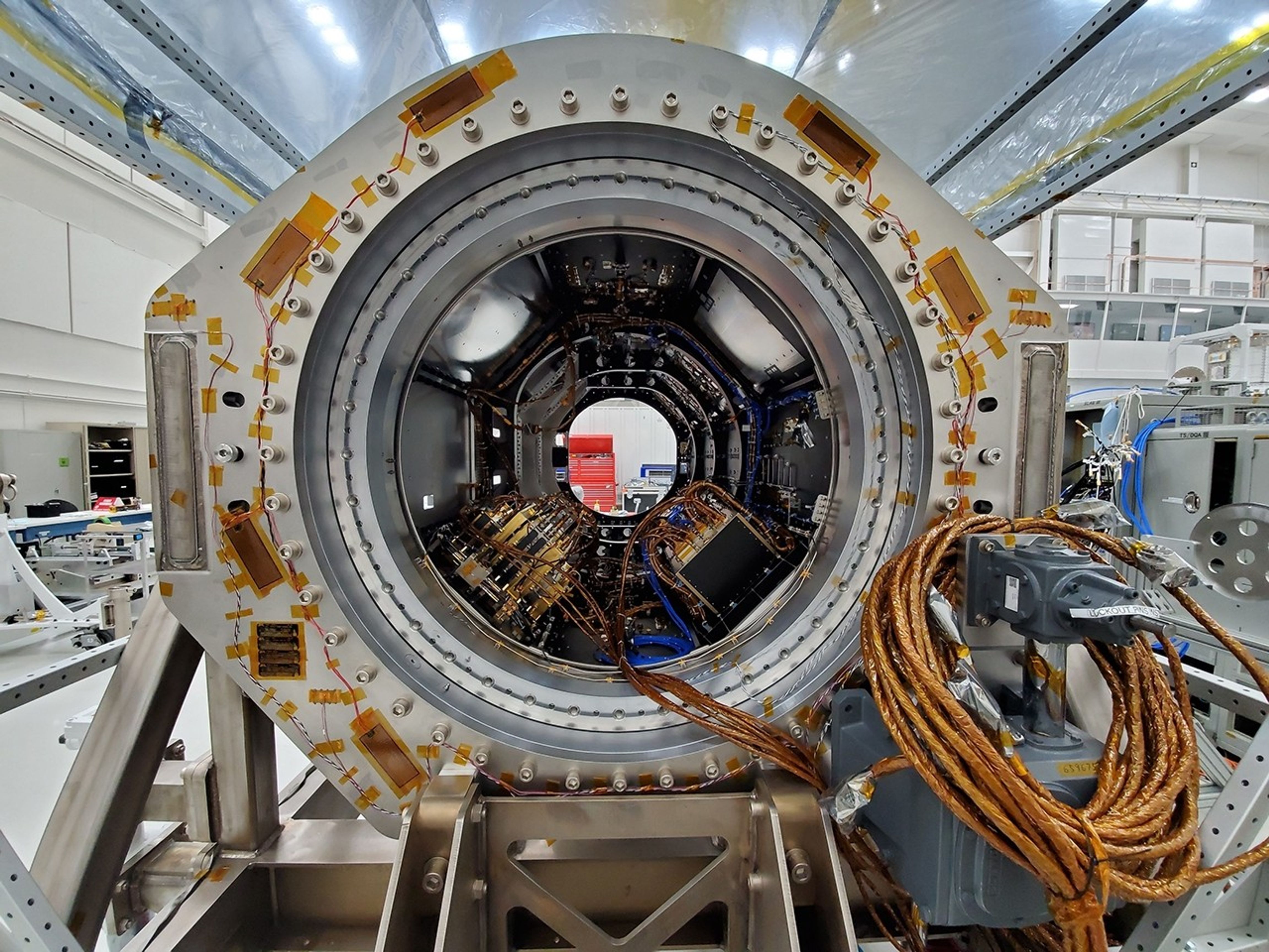a picture of NISAR's integrated L-SAR and S-SAR instruments in the clean room at NASA's Jet Propulsion Laboratory. The instrument is a cylindrical structure with a series of cables running out of the entrance.