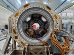 a picture of NISAR's integrated L-SAR and S-SAR instruments in the clean room at NASA's Jet Propulsion Laboratory. The instrument is a cylindrical structure with a series of cables running out of the entrance.