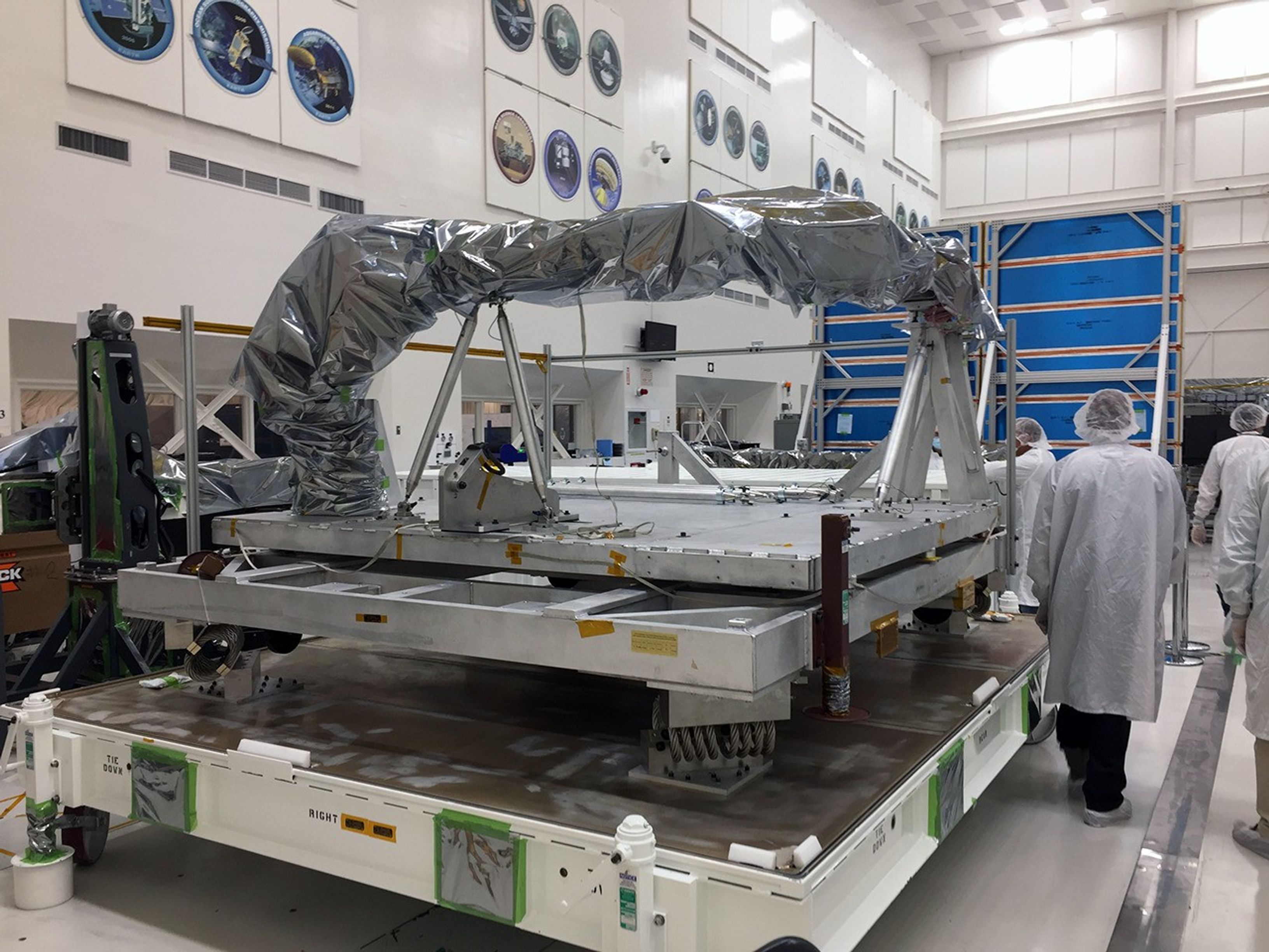 The NISAR upper antenna boom sits on its shipping container in the NASA Jet Propulsion Laboratory clean room. Engineers circle the hardware as its prepared for shipping.