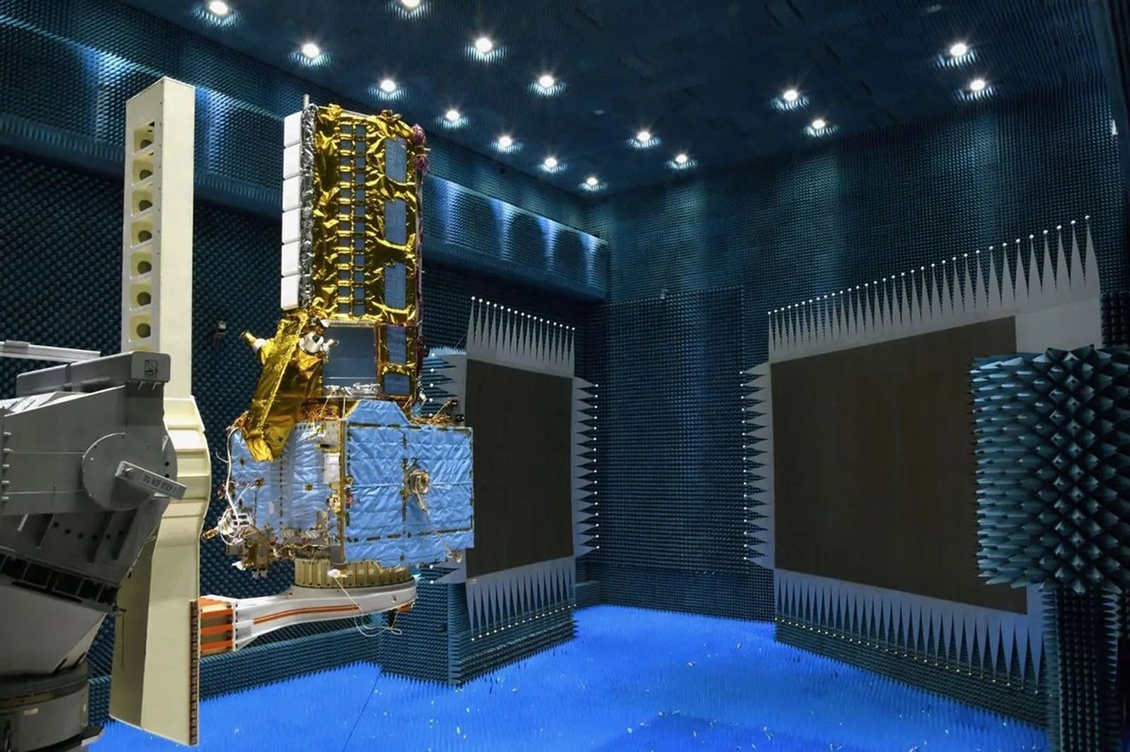 A largely gold covered structure sits on a pedestal on a mechanical arm in a room filled with blue pointed acoustic paneling on the walls and ceiling.