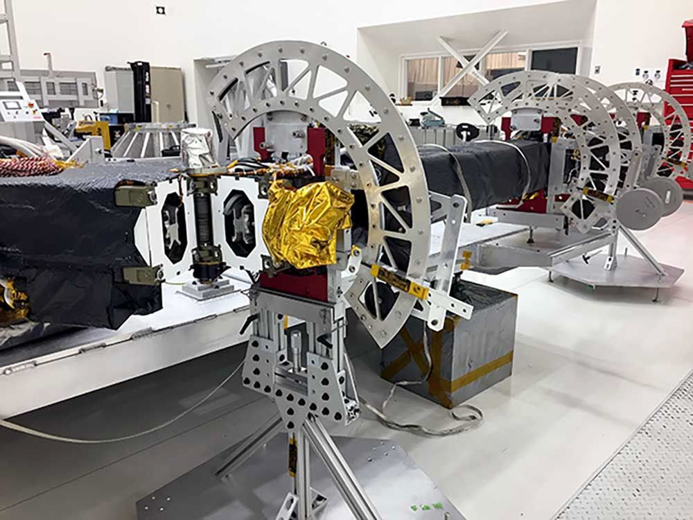 NISAR's upper antenna boom assembly sits in the clean room at NASA's Jet Propulsion Laboratory, ready for assembly. The hardware is a rectangular pole with arching discs of metallic material. Its laying on its side and braced by semicircular metallic braces.