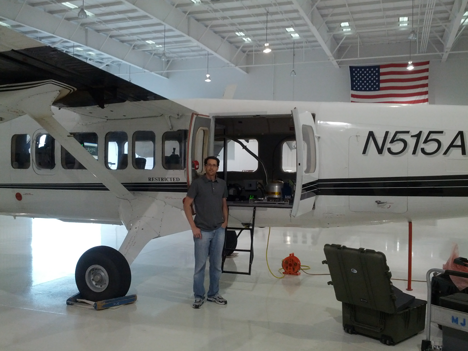 A man in a gray polo shirt and jeans stands next to a white twin-engine propeller aircraft (tail number N515A) inside a hangar. The aircraft's side door is open, revealing equipment inside, including a dome-shaped object. To the right of the aircraft, a green hard case and some orange equipment are on the floor. In the background, a large American flag hangs on the wall, and the hangar ceiling shows exposed beams and lighting fixtures.