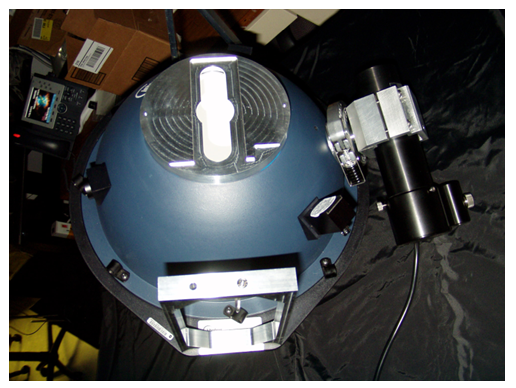 A top-down photograph of a dome-shaped instrument, possibly a radiometer or sensor, with a dark blue surface. Attached to the right side is a cylindrical optical device, colored black and silver, with cables extending from it. The top of the dome features a rectangular white component. The background is a mix of dark fabric and various pieces of equipment, including a small screen displaying a colorful image.