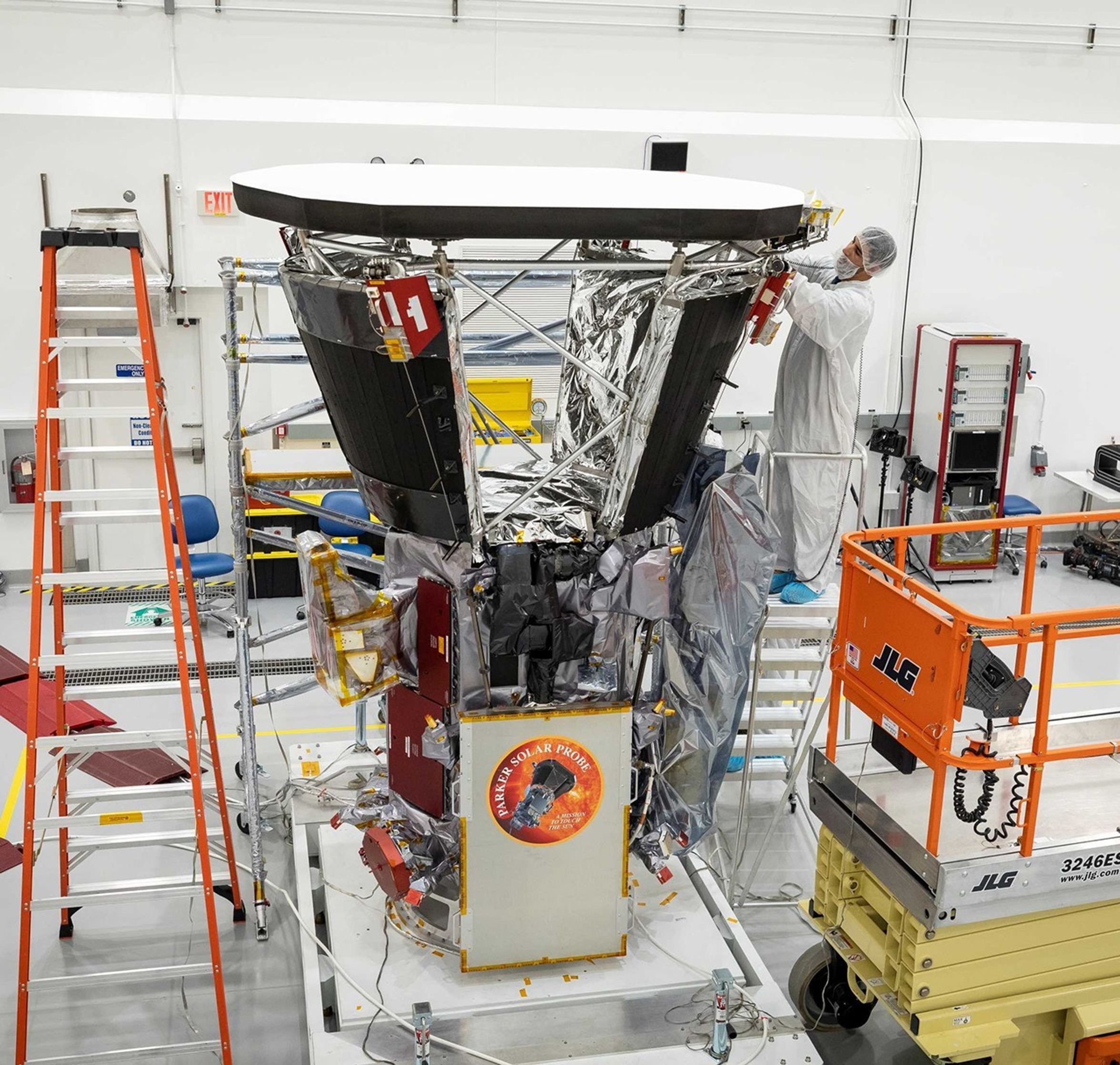 An image of a spacecraft in the center of the photo, with an orange round sticker on the bottom of the spacecraft, identifying it as Parker Solar Probe. A person in a clean-room outfit, right, is installing black thermal shield panels on the spacecraft.