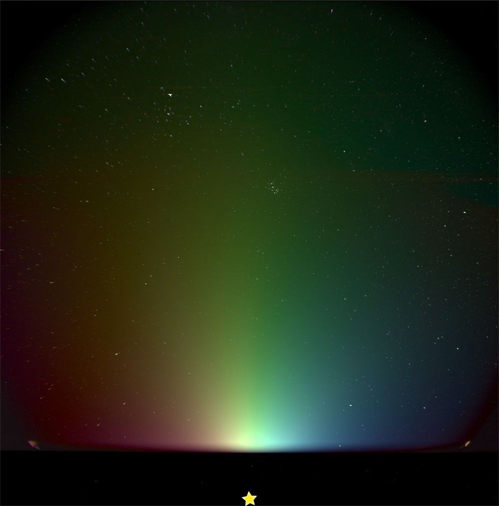 A hazy rainbow-colored glow rises from the bottom of the view, with colors transitioning from red on the far left to green in the center and blue on the far right. The glow is brightest near the bottom center, just above a yellow star symbol, and gradually fades moving upward. The background is black and speckled with stars, including a small cluster to the upper right of center and a larger, V-shaped arrangement of stars in the upper left.