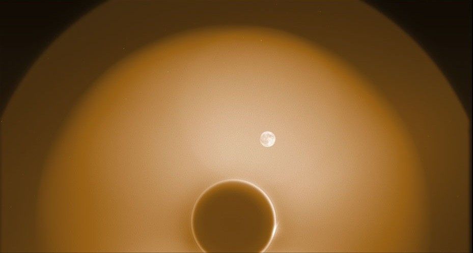 At the bottom, a dark disk is surrounded by a thin bright ring and then by two other larger, circular glows, with the inner circular glow larger and brighter than the outer one. The Moon appears to the upper right of the dark disk, inside the larger and brighter, innermost circular glow.