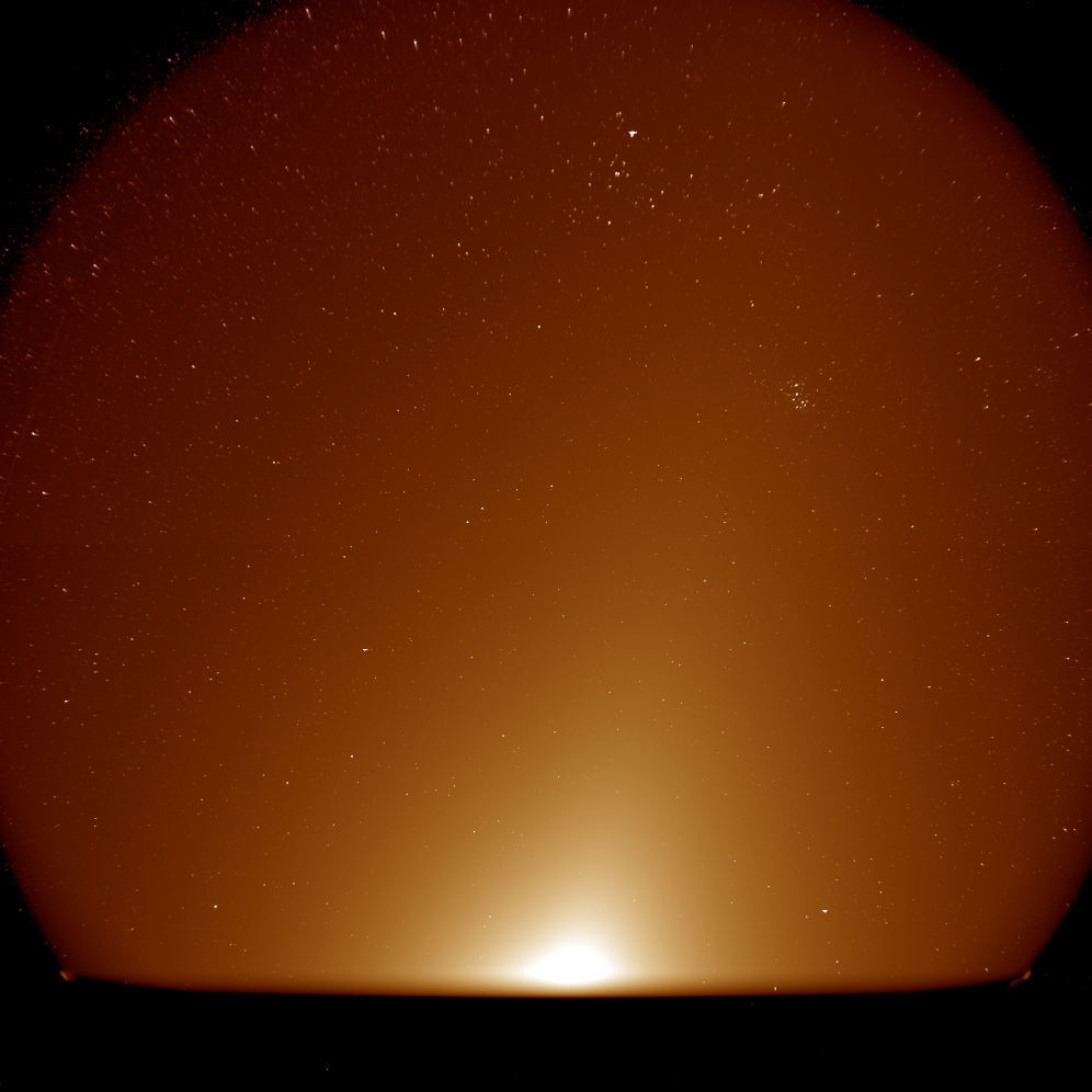A hazy orange glow rises from the bottom of the view against a background of stars. The glow is brightest near the bottom center and gradually fades moving upward, but with a concentrated beam angled toward the upper right. A small star cluster appears in the upper right and a larger, V-shaped arrangement of stars appears near the top.