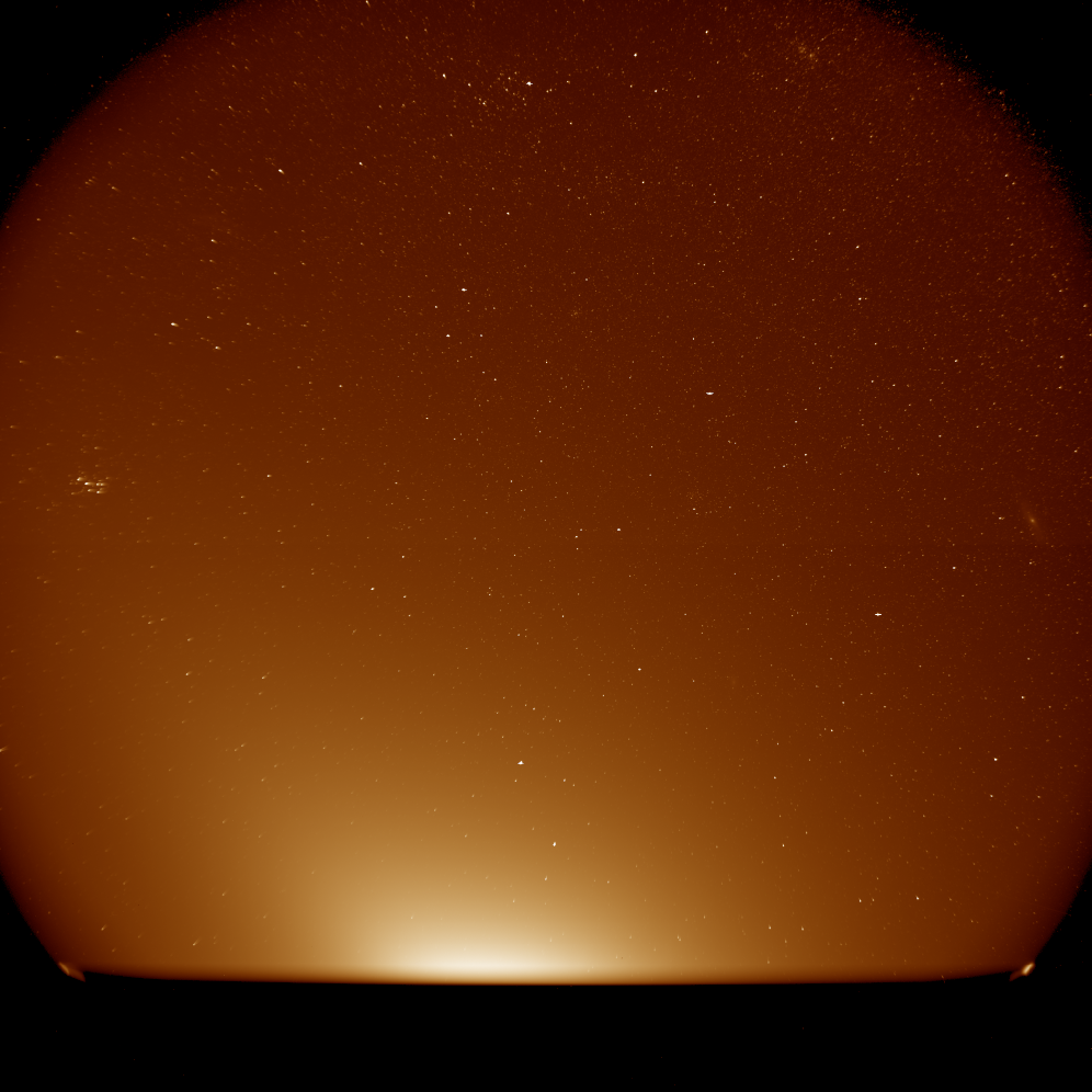 A hazy orange glow rises from the bottom of the view against a background of stars. The glow is brightest near the bottom center and gradually fades moving upward, but with a concentrated beam angled toward the upper left. A small star cluster appears in the upper left, and a small, extended hazy object appears in the upper right.