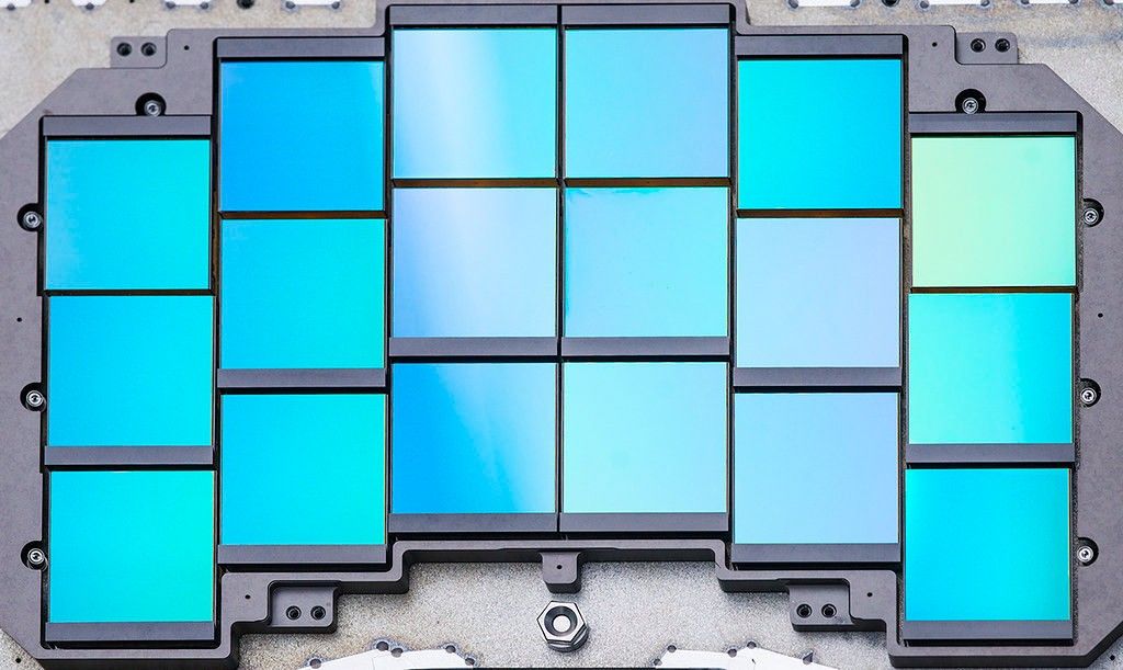 Three stacked, arching rows of six squares each, all embedded in steely gray metal hardware. The squares are shades of blue-green. Together, they resemble a space invader.