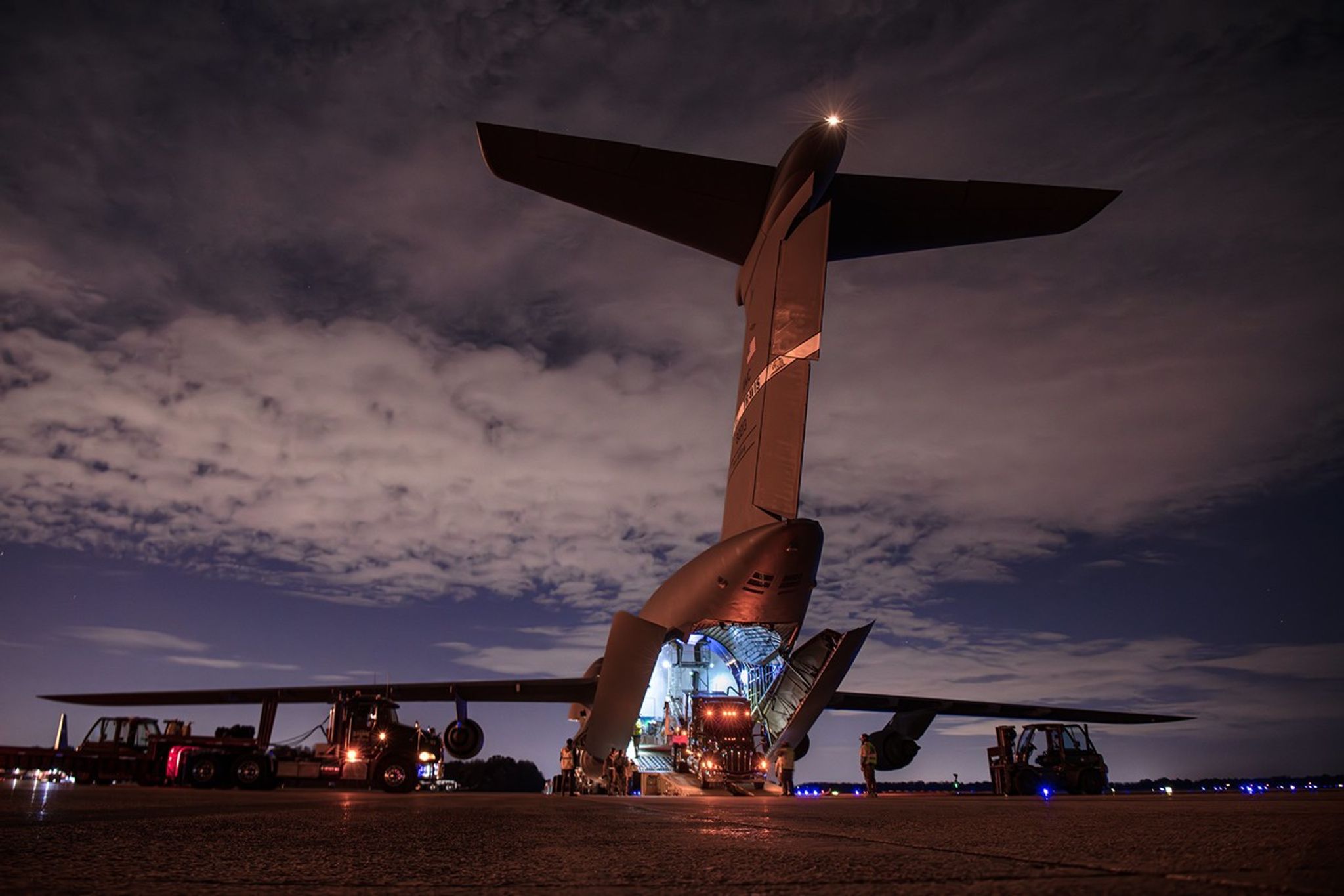 A large, dimly lit plane is seen from behind at night. The back end is open and lights are on inside. Something large is moving down a ramp to exit the airplane.