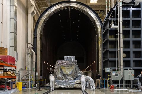 A wide shot of a silvery piece of space hardware about the size of a car sitting on a silver…