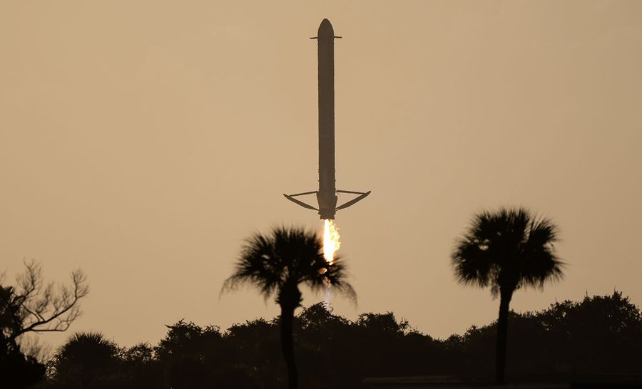 A rocket booster descends vertically with its landing engines firing, silhouetted against a golden sky. The bright plume from the engine shines through the tops of nearby palm trees, which frame the scene in the foreground.