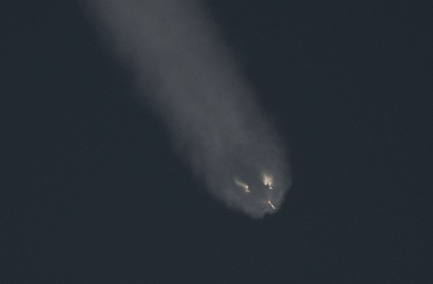 A rocket stage descends against a dark sky, trailing a wide plume of vapor. Several bright engine flames are visible at the bottom of the plume, creating small points of light as the booster returns through the atmosphere.