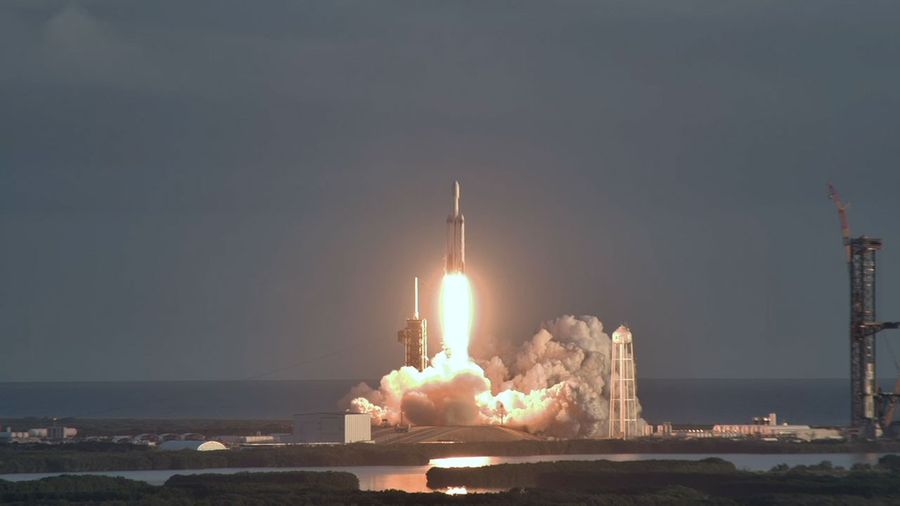 A heavy‑lift rocket rises from the launch pad, its engines producing a bright plume of fire and thick clouds of exhaust. The vehicle ascends into a dark sky above the coastline, with launch towers and ground facilities visible below.