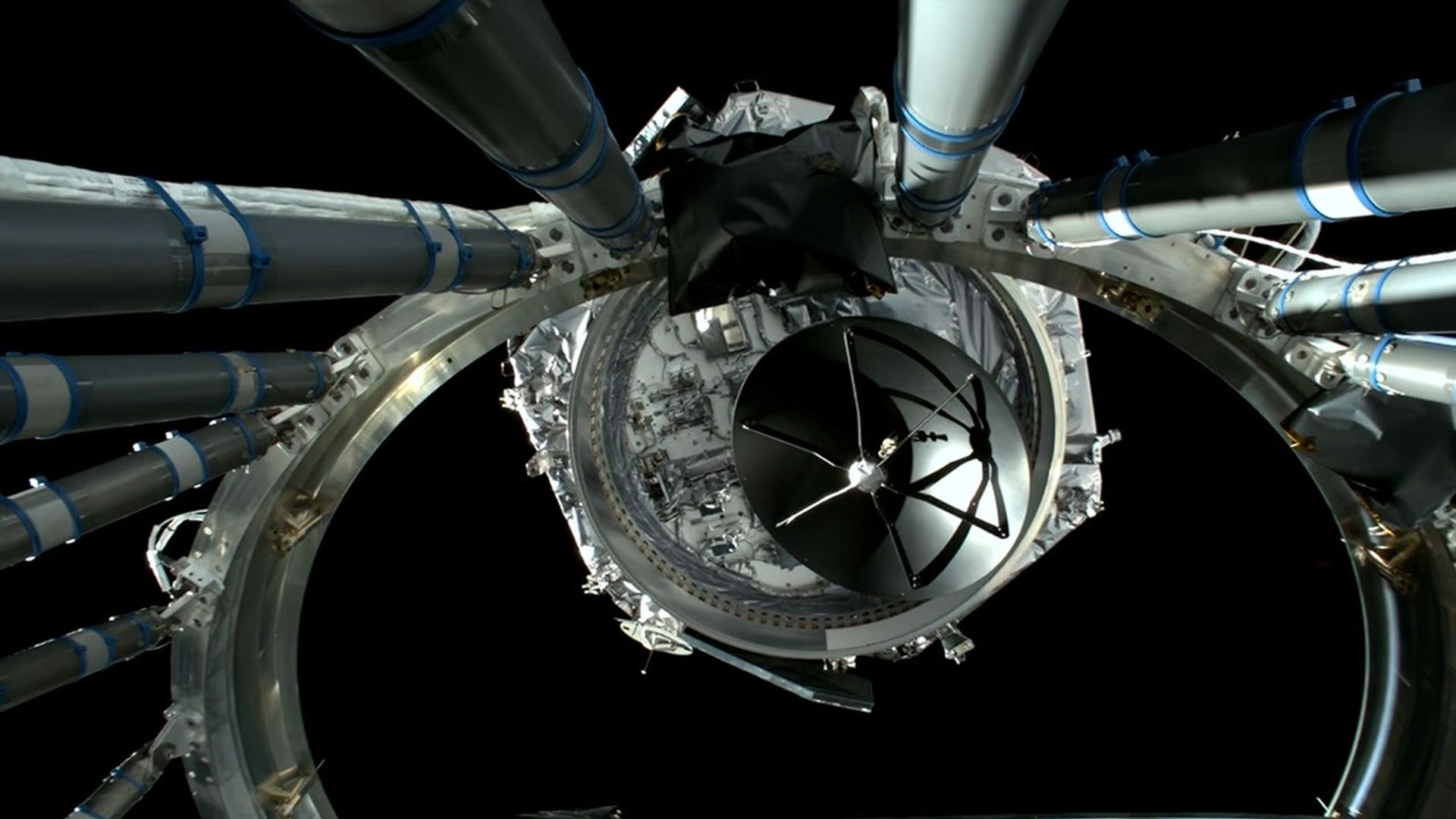 View from the rocket’s upper stage showing the spacecraft emerging after separation, framed by structural support arms and metallic wiring against the blackness of space.