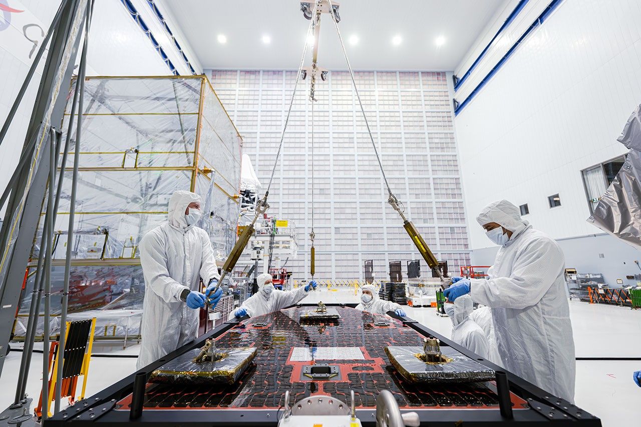 Four technicians in clean suits hook a solar panel up to a crane lift.