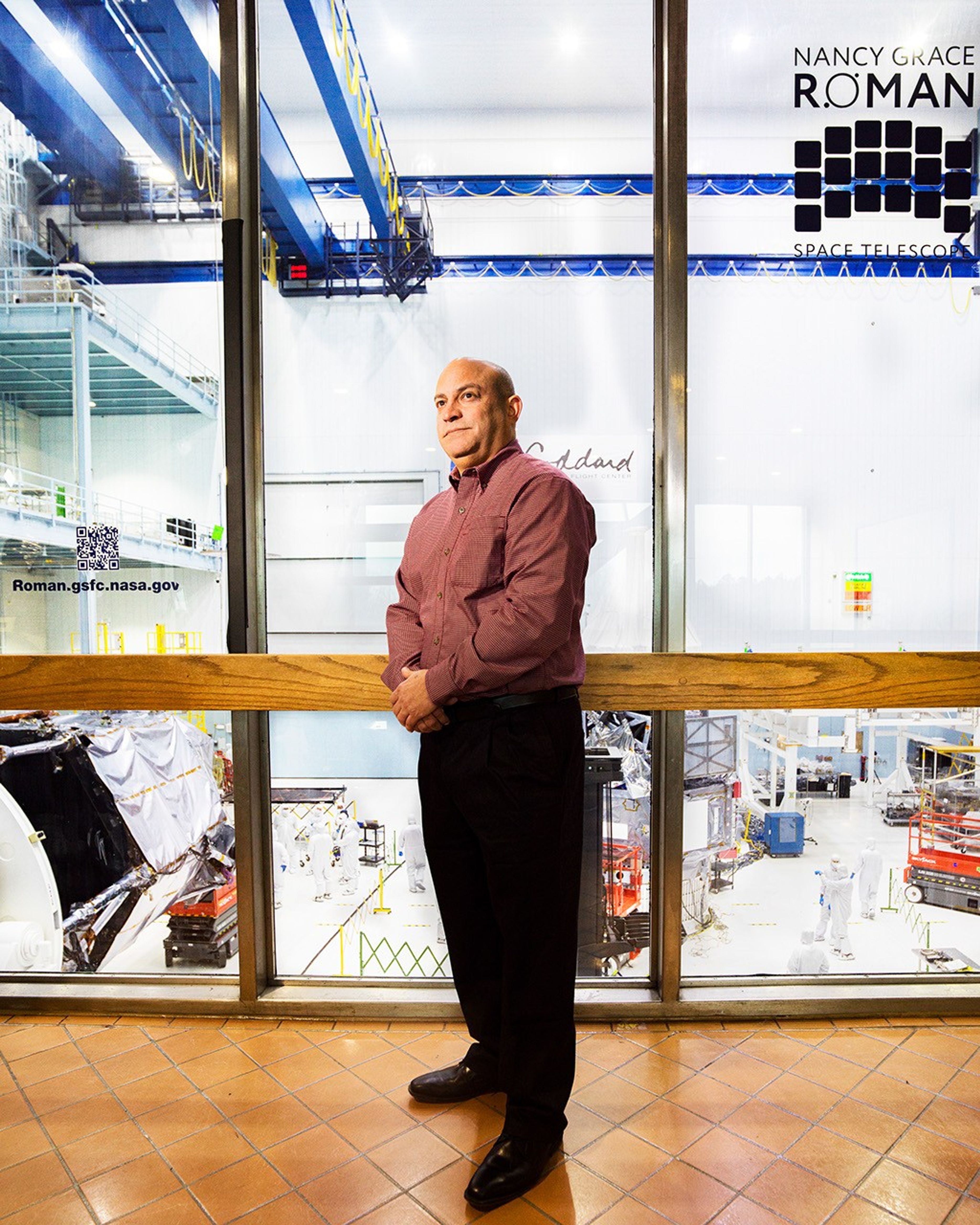 A man stands in front of a floor-to-ceiling window, through which major space hardware components are visible.