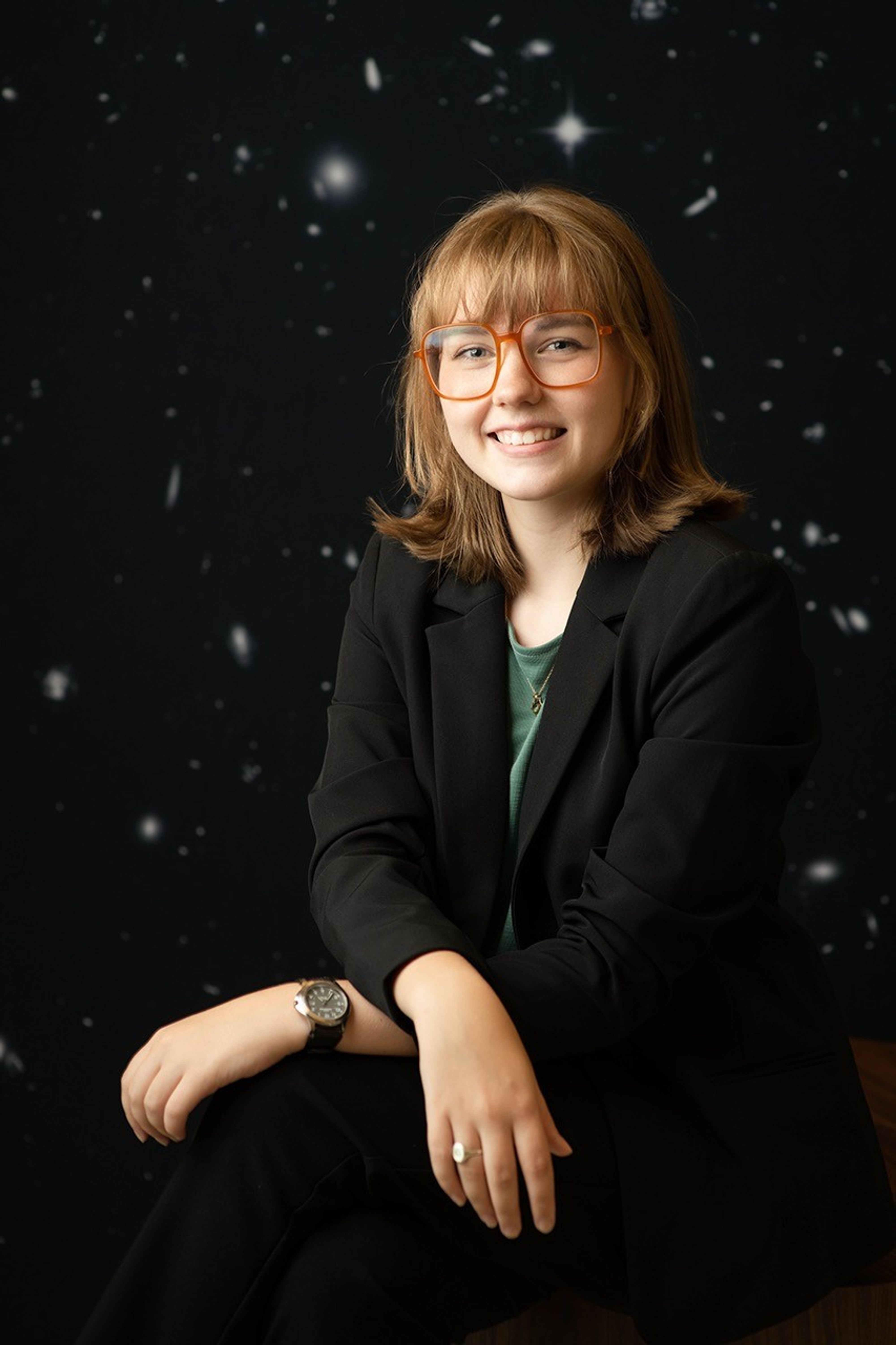 A smiling young woman with red glasses and shoulder length dark blonde hair sits in front of a backdrop of tiny galaxies speckling a black background.
