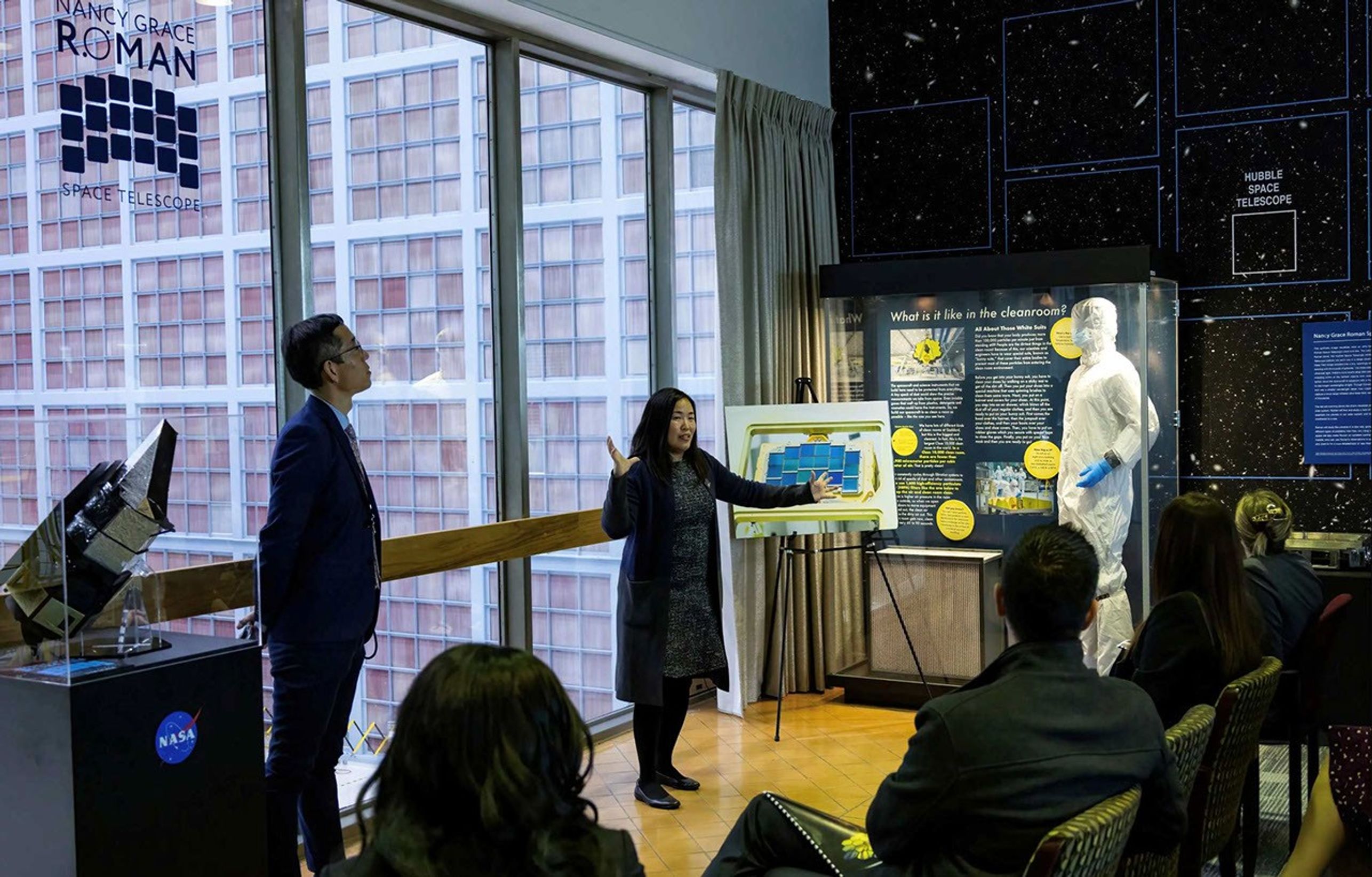 A woman standing with her arms outstretched addresses a small crowd in a dimly lit room. Through a floor-to-ceiling glass window behind her, a large wall is covered in a grid of small, pale pink squares. Inside the room to the woman’s left, a man stands beside a small model of the Nancy Grace Roman Space Telescope, which is encased in glass. To the woman’s right, a large photo of Roman’s detectors is propped up on an easel. In a large glass case next to it, a life-size mannequin is dressed in clean room garb: a head-to-toe white suit, white face covering, and blue gloves.