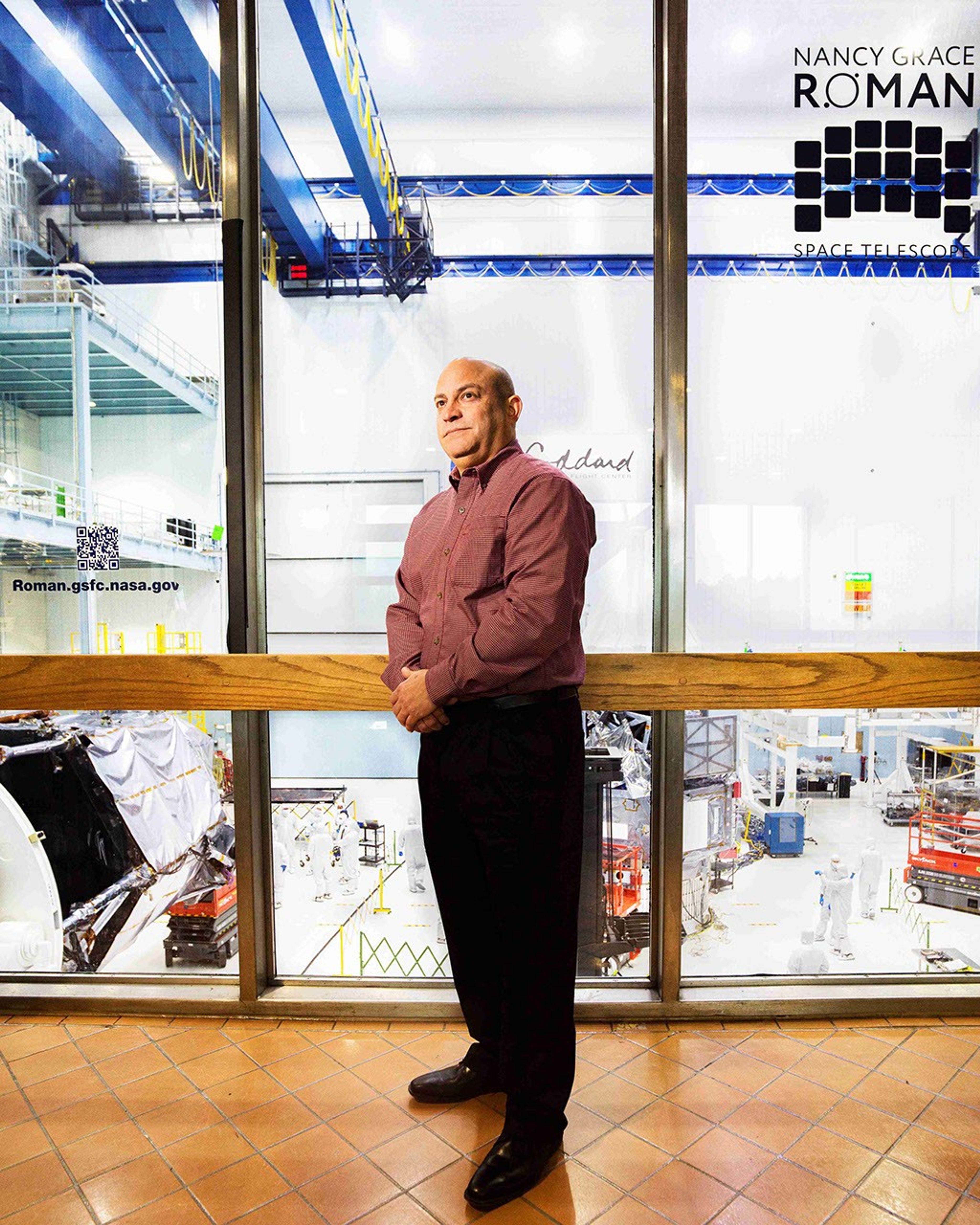 A man stands in front of a floor-to-ceiling window, through which major space hardware components are visible.