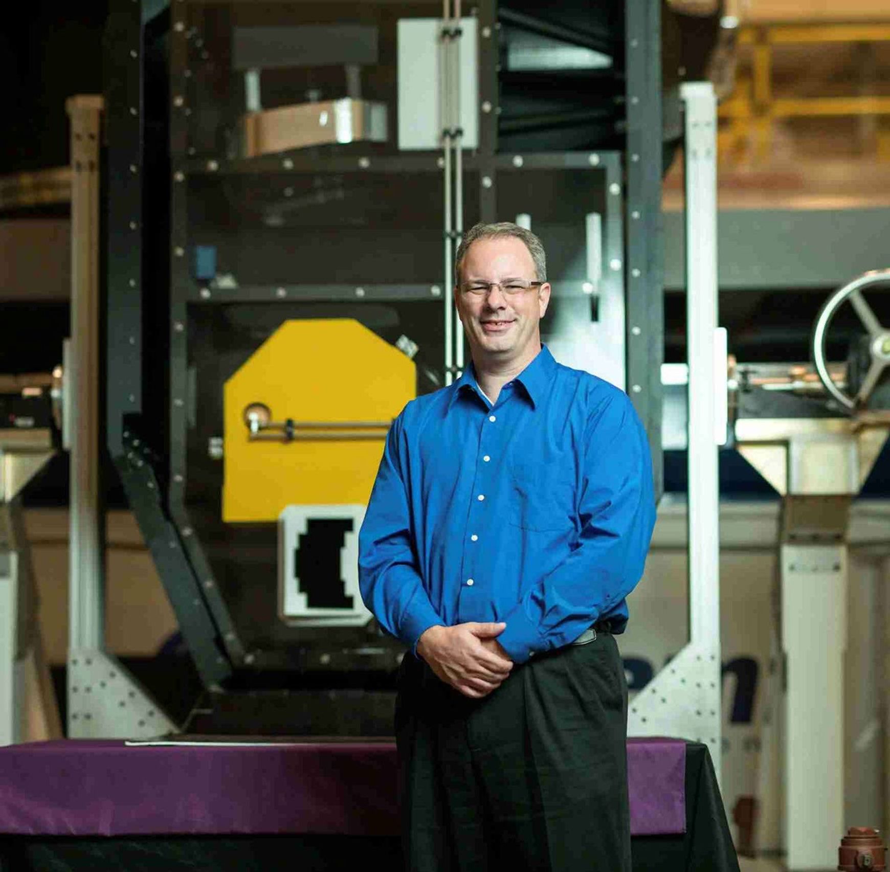 A smiling man wearing glasses, a long sleeve blue button up shirt, and black pants stands with his arms crossed. Behind him in a large clear case with black and metallic edges there is a gold colored pane.