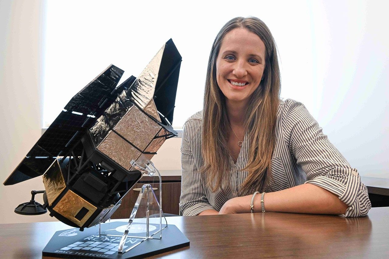 A smiling woman sits at a desk. On the desk beside her there is a model of the Nancy Grace Roman Space Telescope.