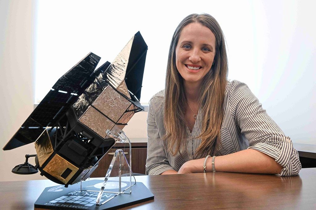 A smiling woman sits at a desk. On the desk beside her there is a model of the Nancy Grace Roman Space Telescope.