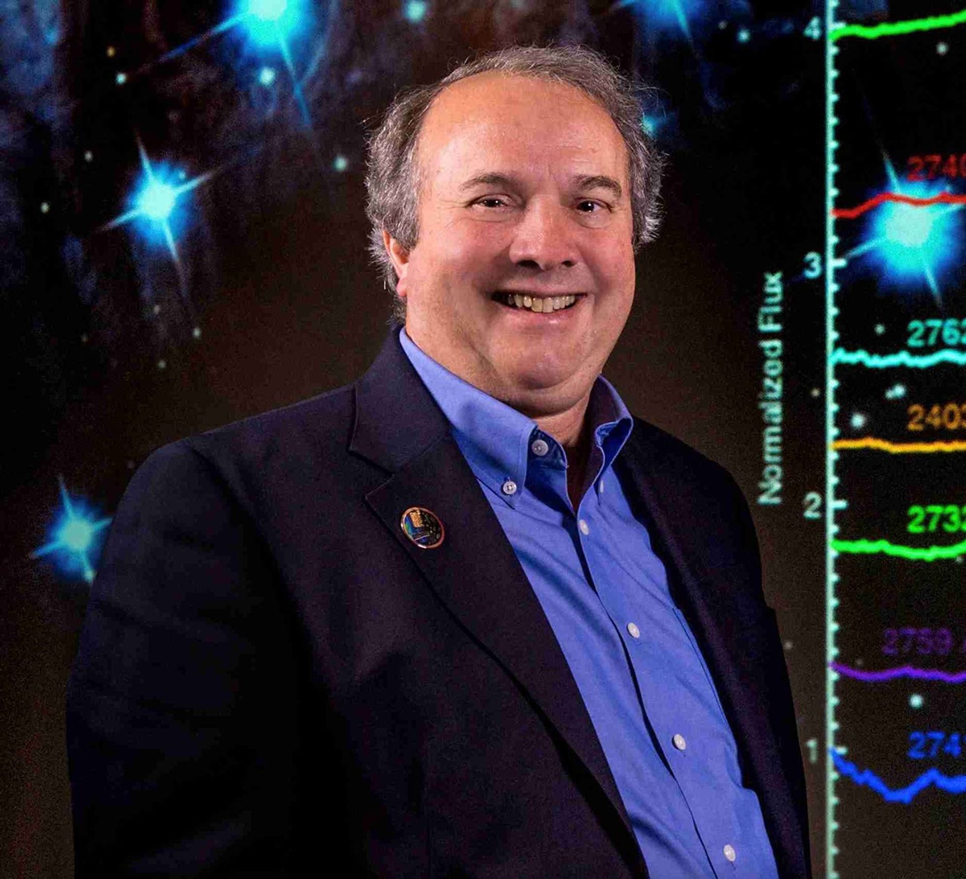 A man in a suit stands in front of a background with bright blue stars with splinters of starlight coming out of them like crosses at the left and above him. Behind and at his right, there’s a vertical ruler labeled “Normalized Flux.” Brightly colored squiggly lines extend from different heights on the ruler to the right, off screen.