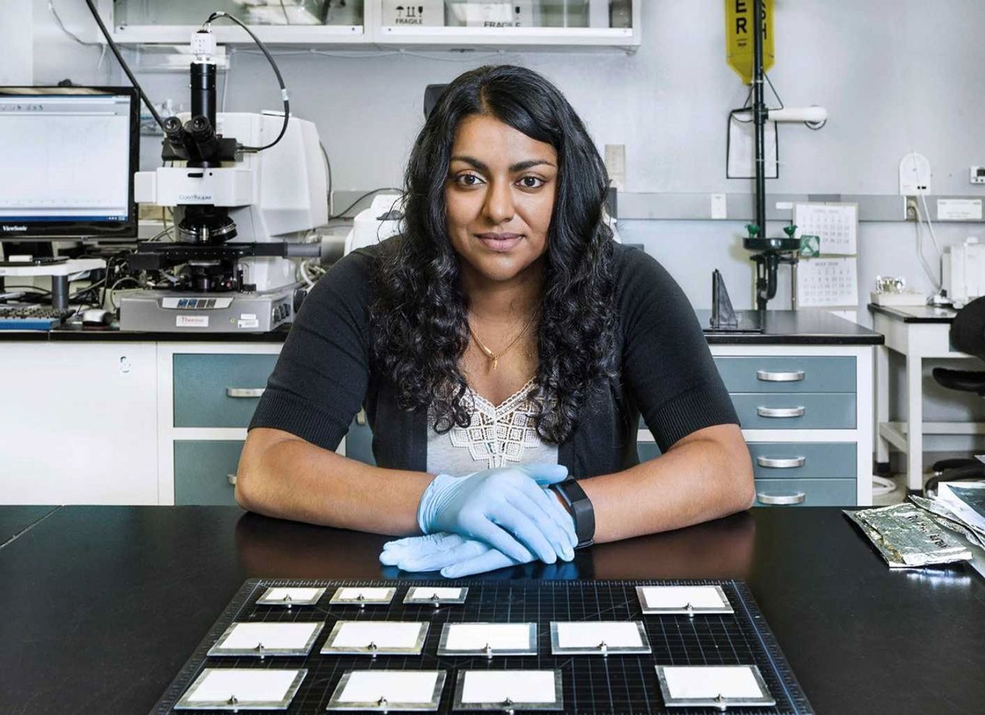 A woman sits at a black table with gloved hands on the tabletop. Laboratory equipment is in the background.