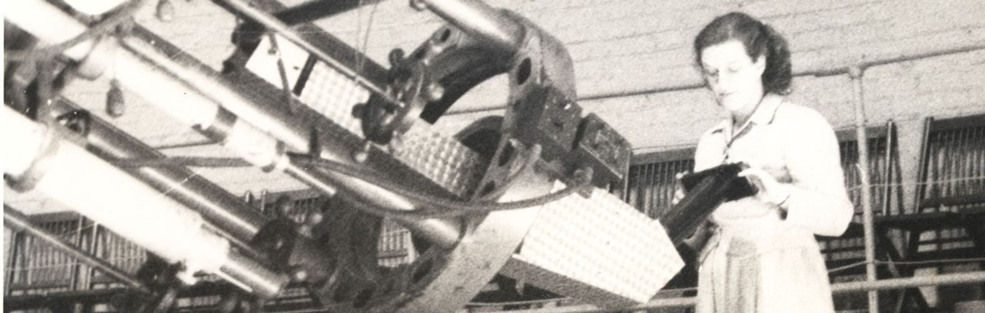 The 1948 image shows Astronomer Nancy Grace Roman, a woman scientist inspecting the spectrograph attachment of the Yerkes Observatory telescope in Williams Bay, Wisconsin. The telescope is a metallic structure with several aligned tubes and a circular component. In the background is a row of folding chairs.