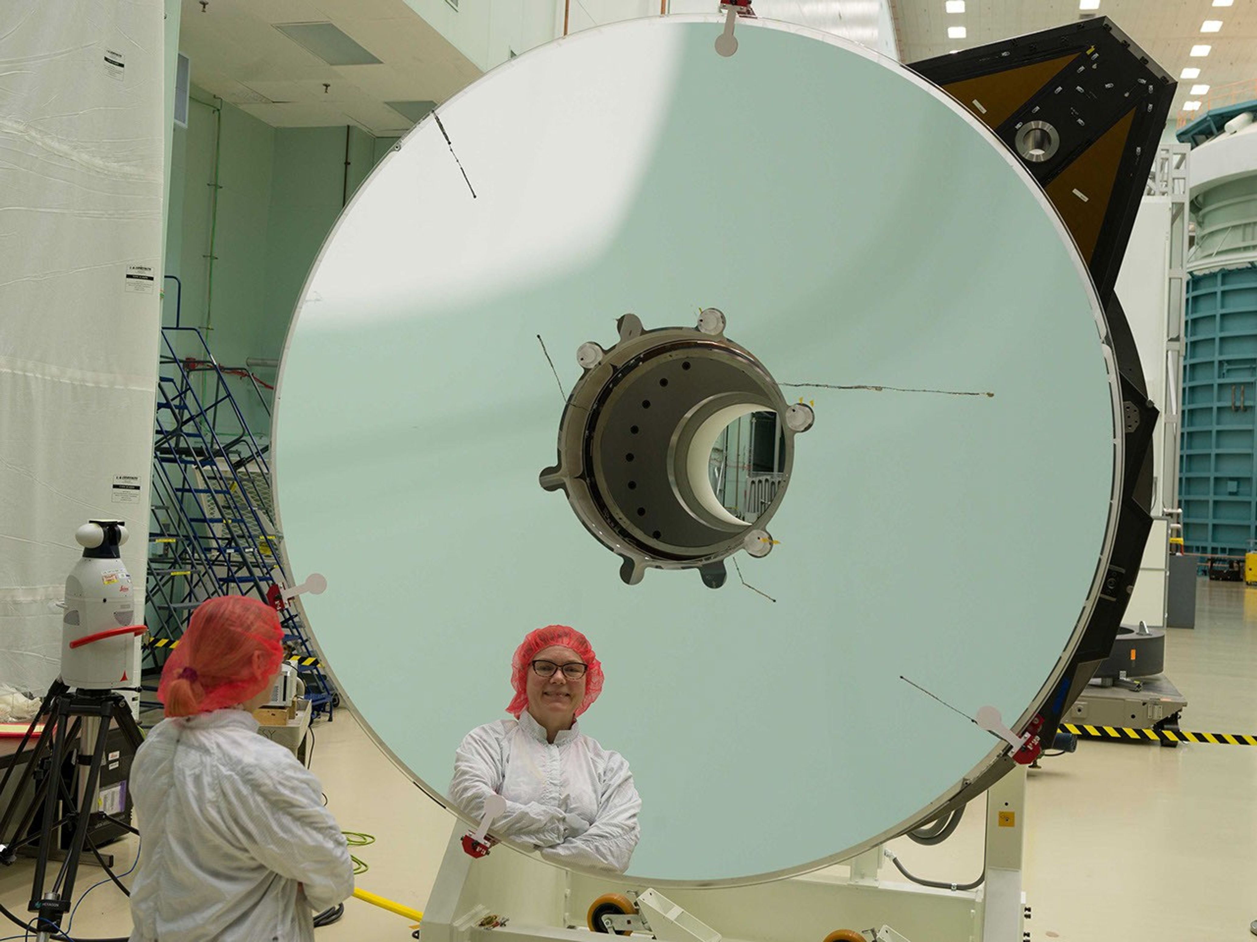 A woman wearing a white jumpsuit, red hairnet, and black framed glasses stands in front of a large circular mirror with a smaller circle cut out in the middle. She faces away from the camera and her reflection is shown in the mirror.