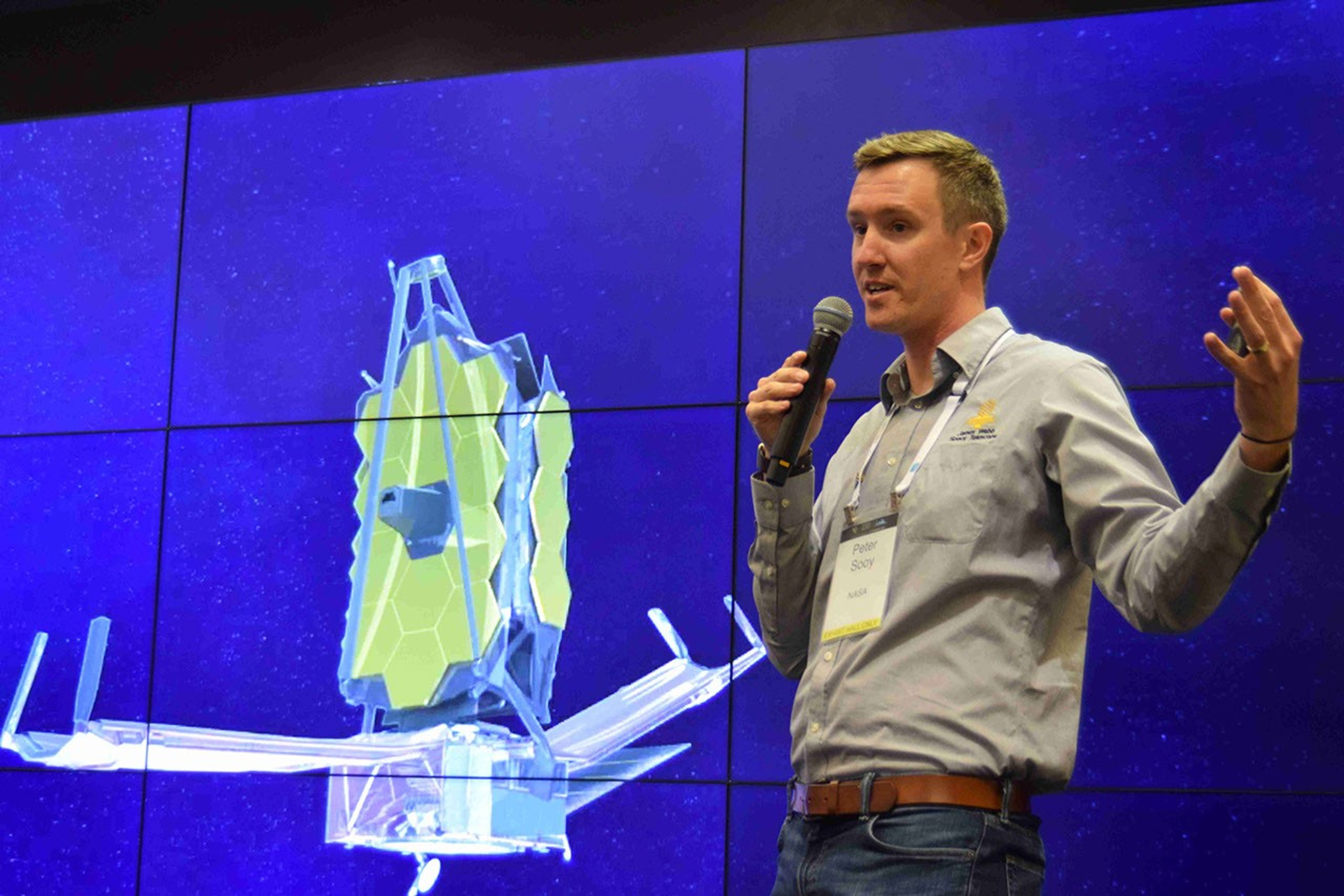 A man with dark blonde hair speaks into a microphone in front of a backdrop. The backdrop is deep blue and features an image of the James Webb Space Telescope, which has a silvery base with �wings� that extend outward from each side and a golden honeycomb-like mirror that protrudes from the top.