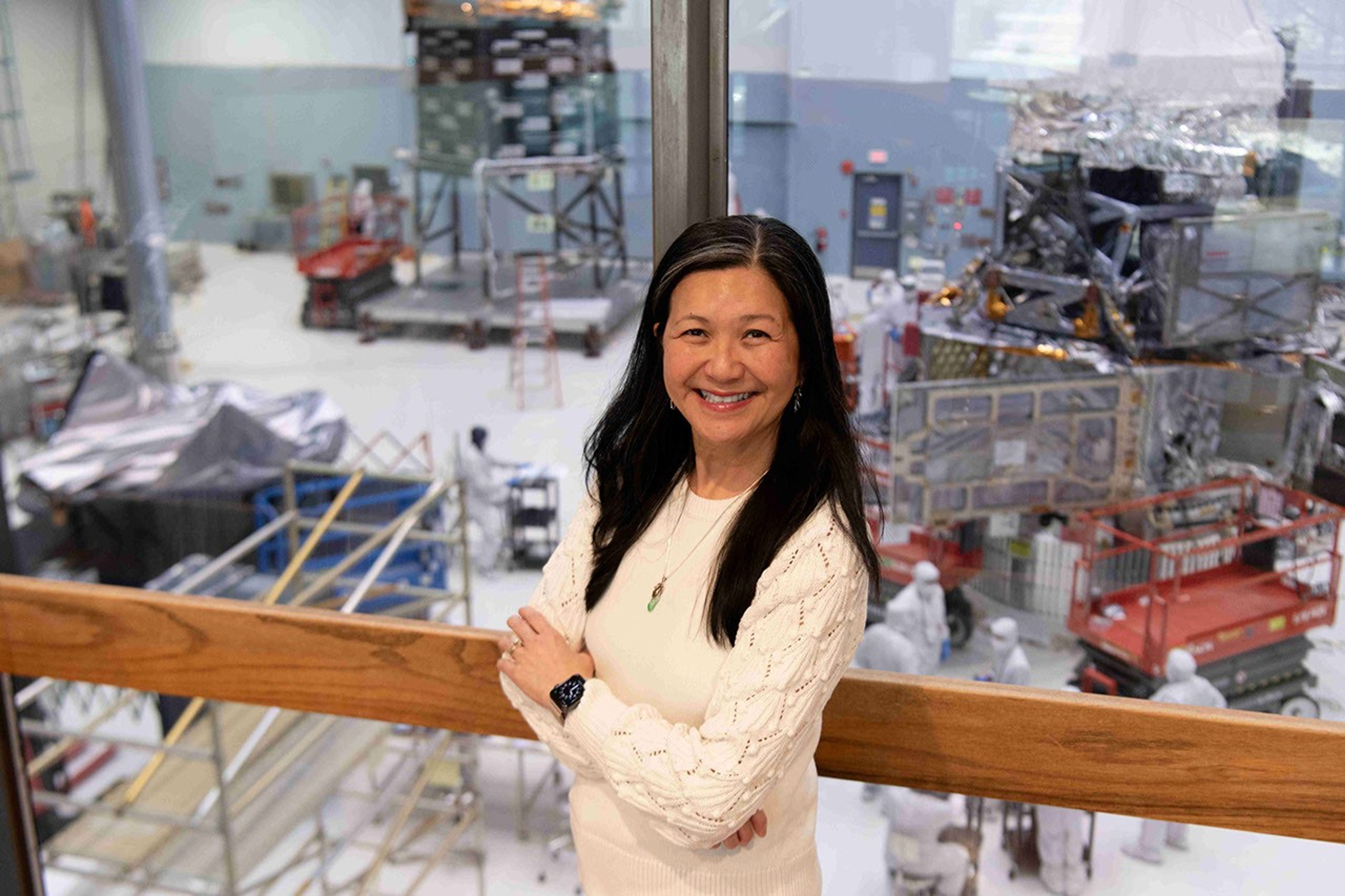A smiling woman stands in front of a floor-to-ceiling window, through which many large space hardware components are visible.