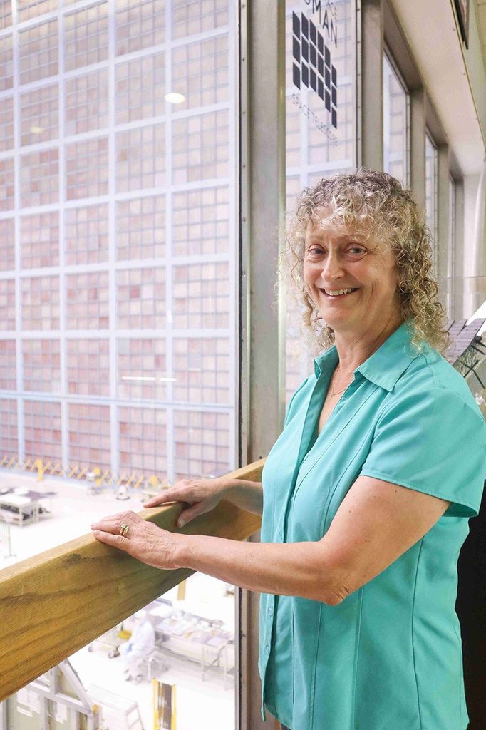 An image of a woman with curly hair in a sea foam green shirt stands with her hands on a railing in front of a floor to ceiling window. Through the window, there’s a view into a large bright room with a grid of pale pink squares on the far wall, which is multiple stories high.