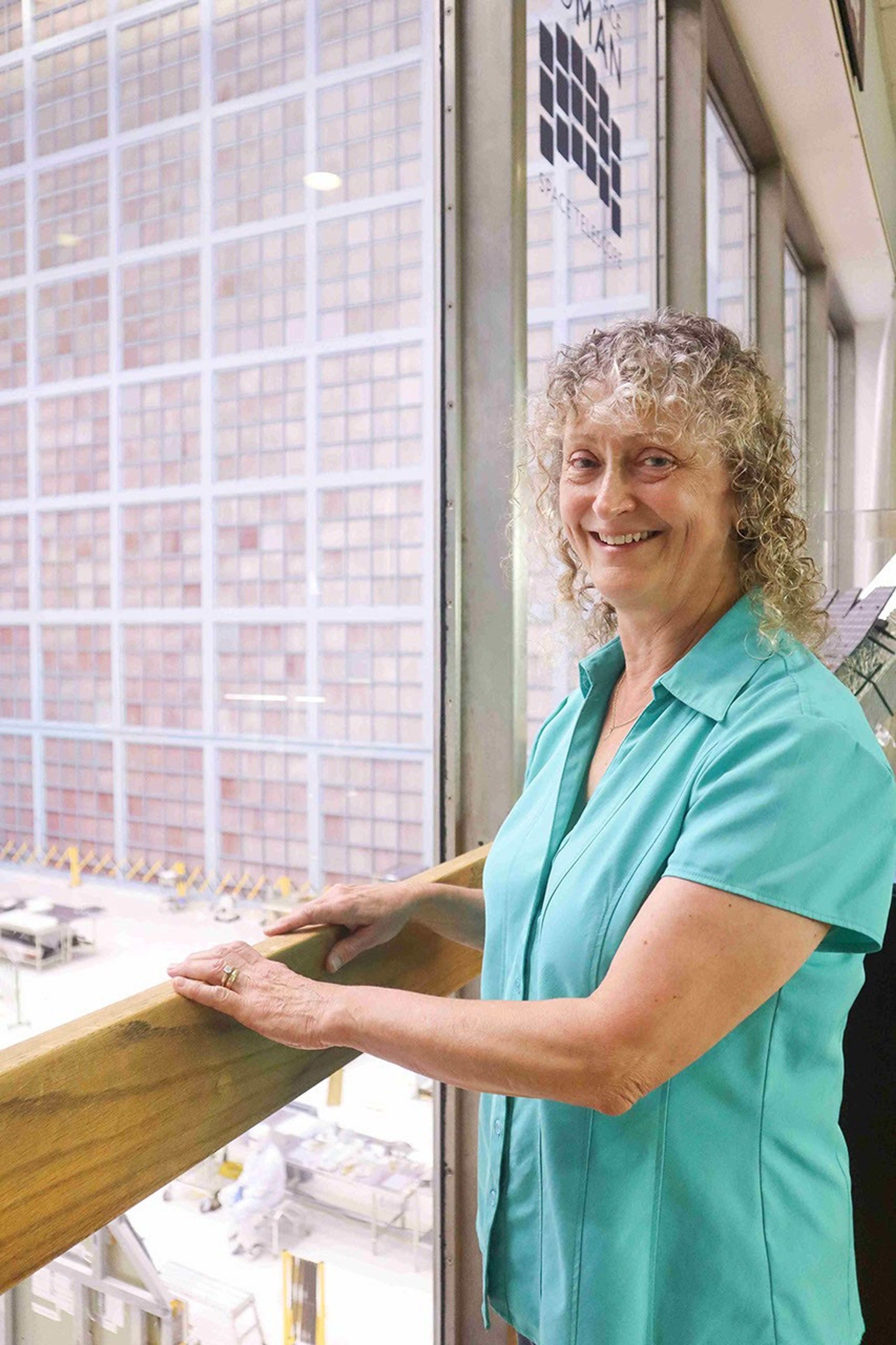 An image of a woman with curly hair in a sea foam green shirt stands with her hands on a railing in front of a floor to ceiling window. Through the window, there’s a view into a large bright room with a grid of pale pink squares on the far wall, which is multiple stories high.