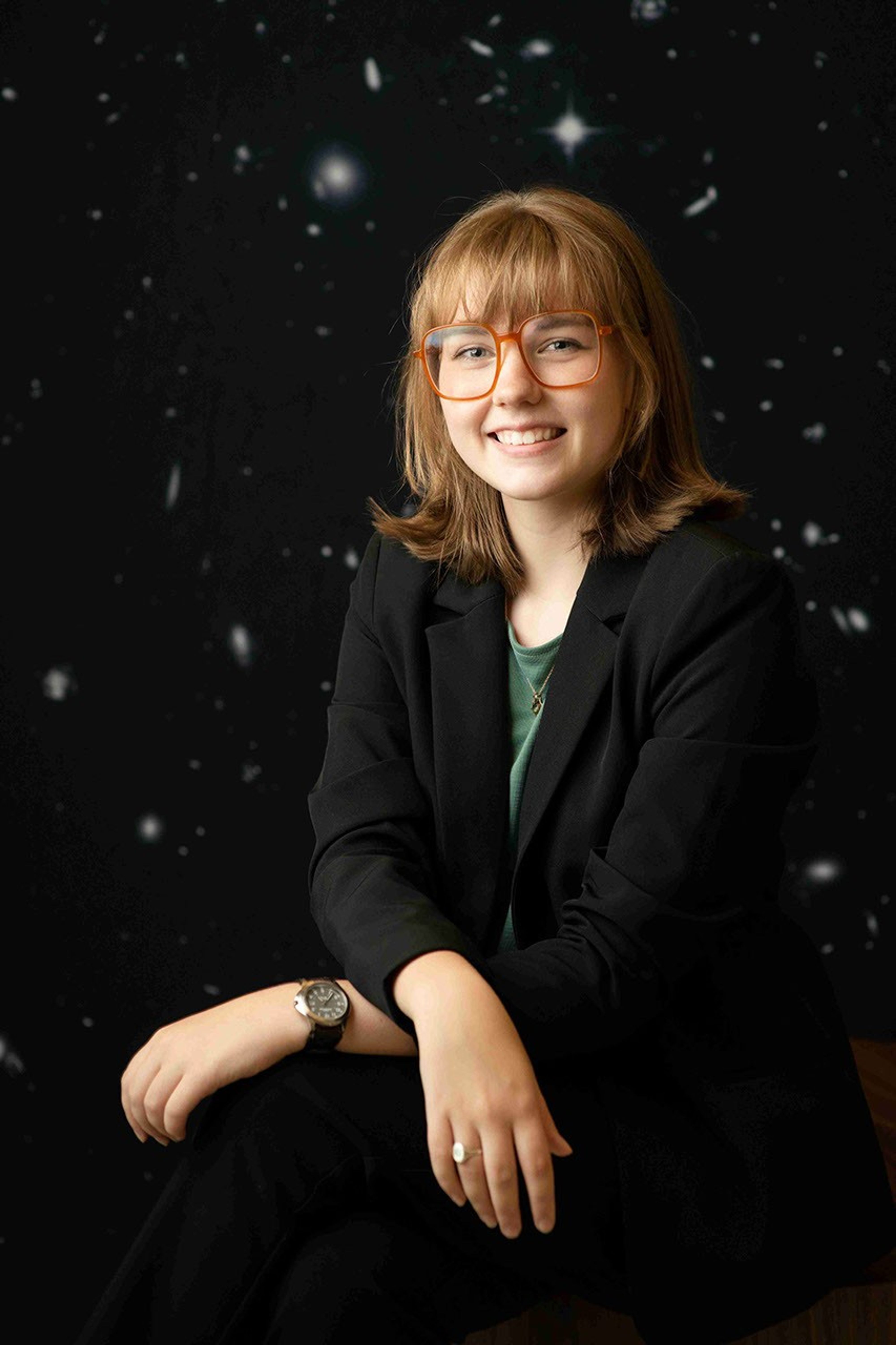 A smiling young woman with red glasses and shoulder length dark blonde hair sits in front of a backdrop of tiny galaxies speckling a black background.