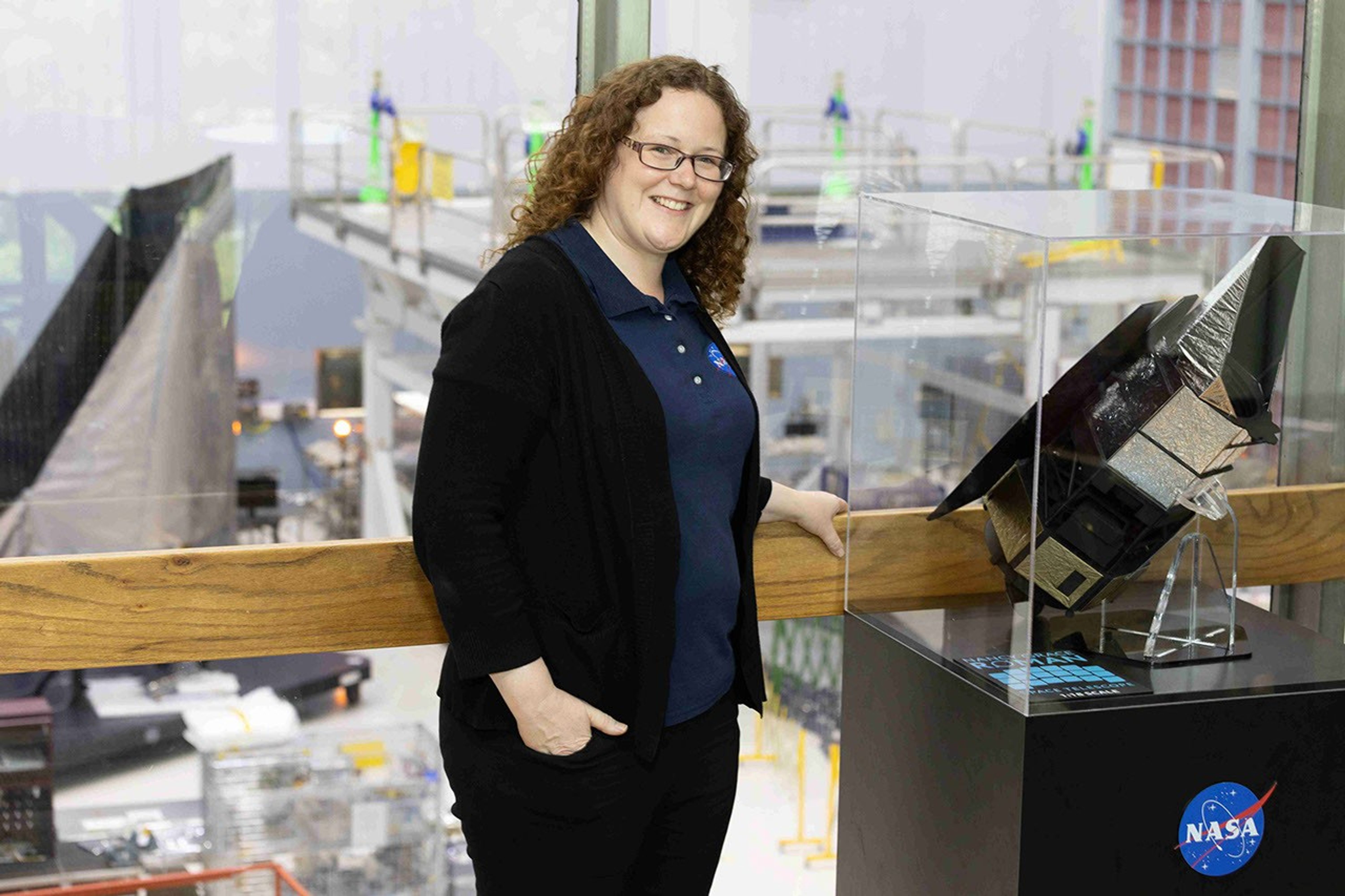A woman with curly red hair stands to the left of a telescope model in a glass case. Behind her (visible through a floor to ceiling glass window), a full-scale version of the same telescope is under construction.