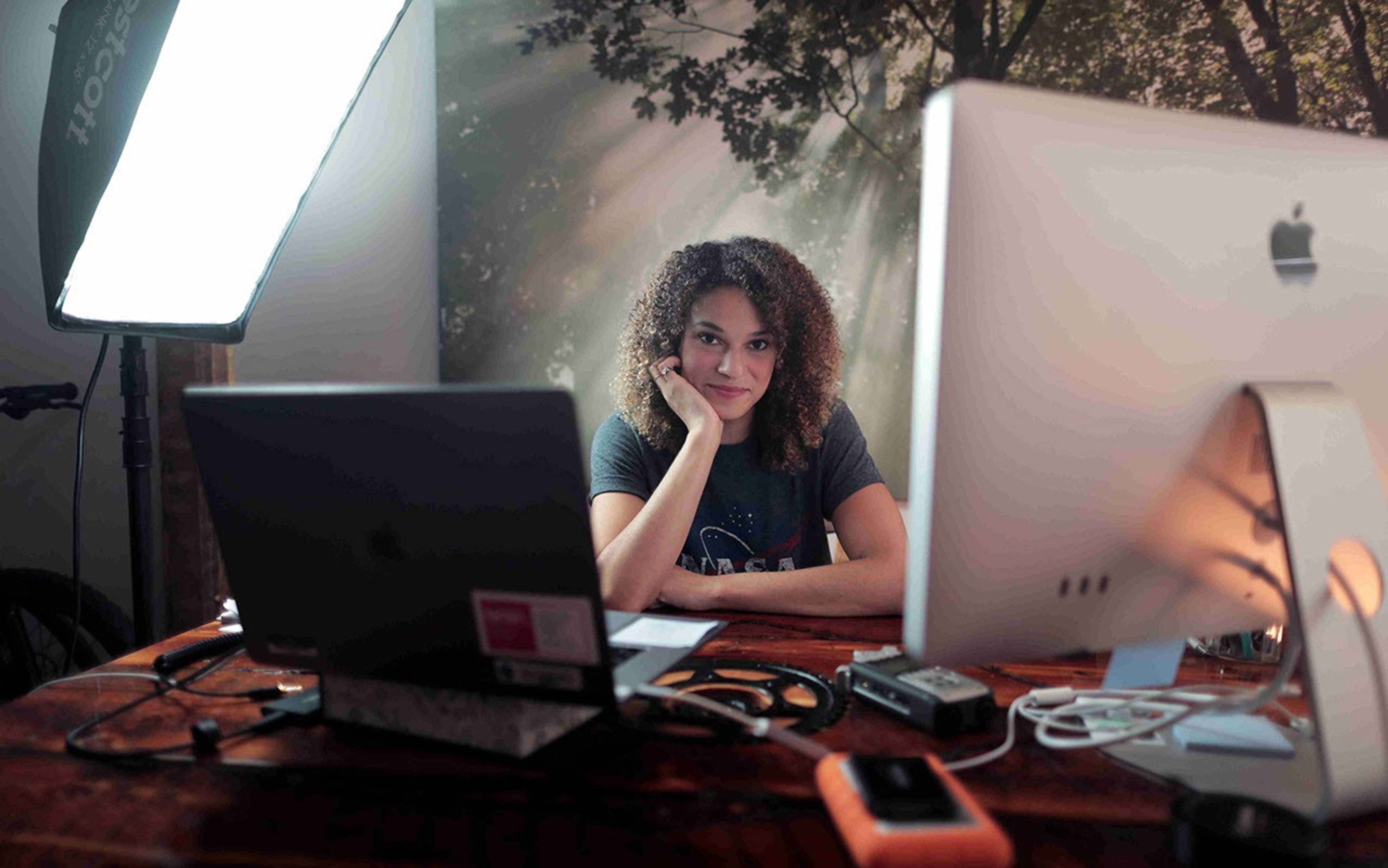 A woman sits at a desk with two computer monitors in the foreground.